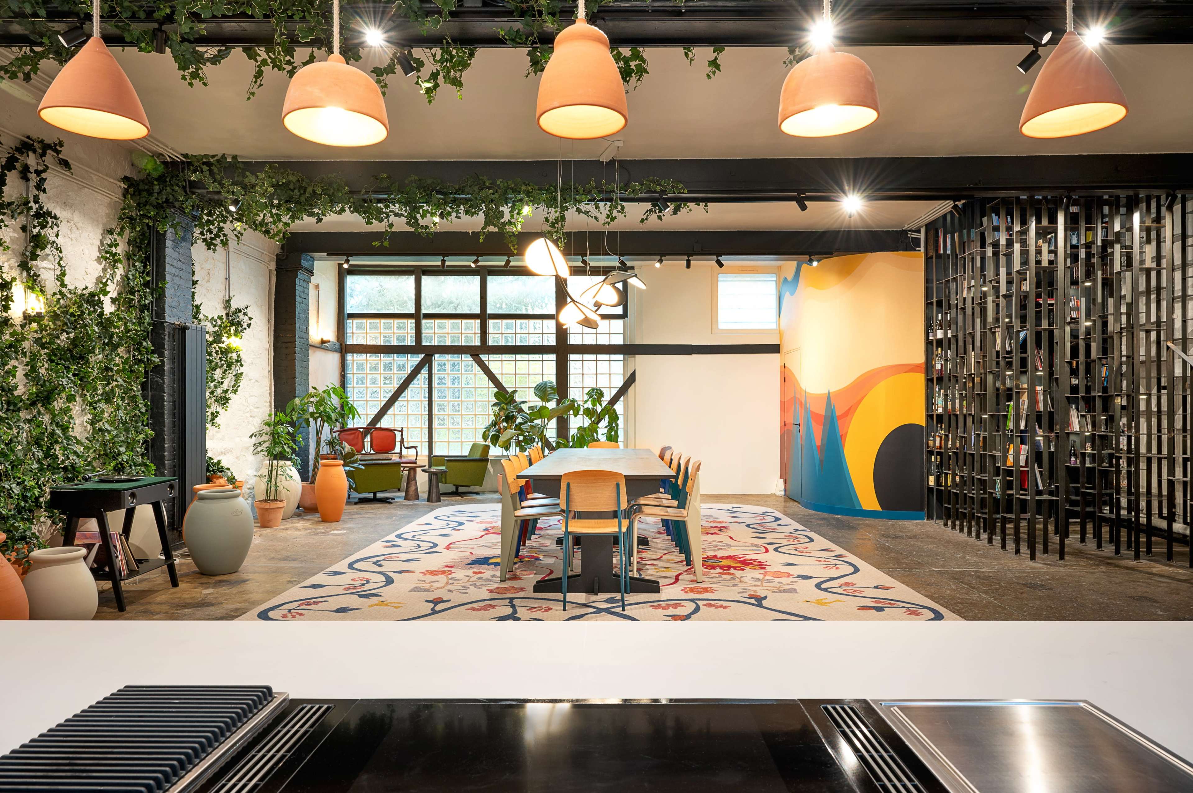 The image shows a modern office space featuring a long dining table surrounded by chairs, with greenery on the walls, colorful artwork, and various light fixtures hanging from the ceiling.