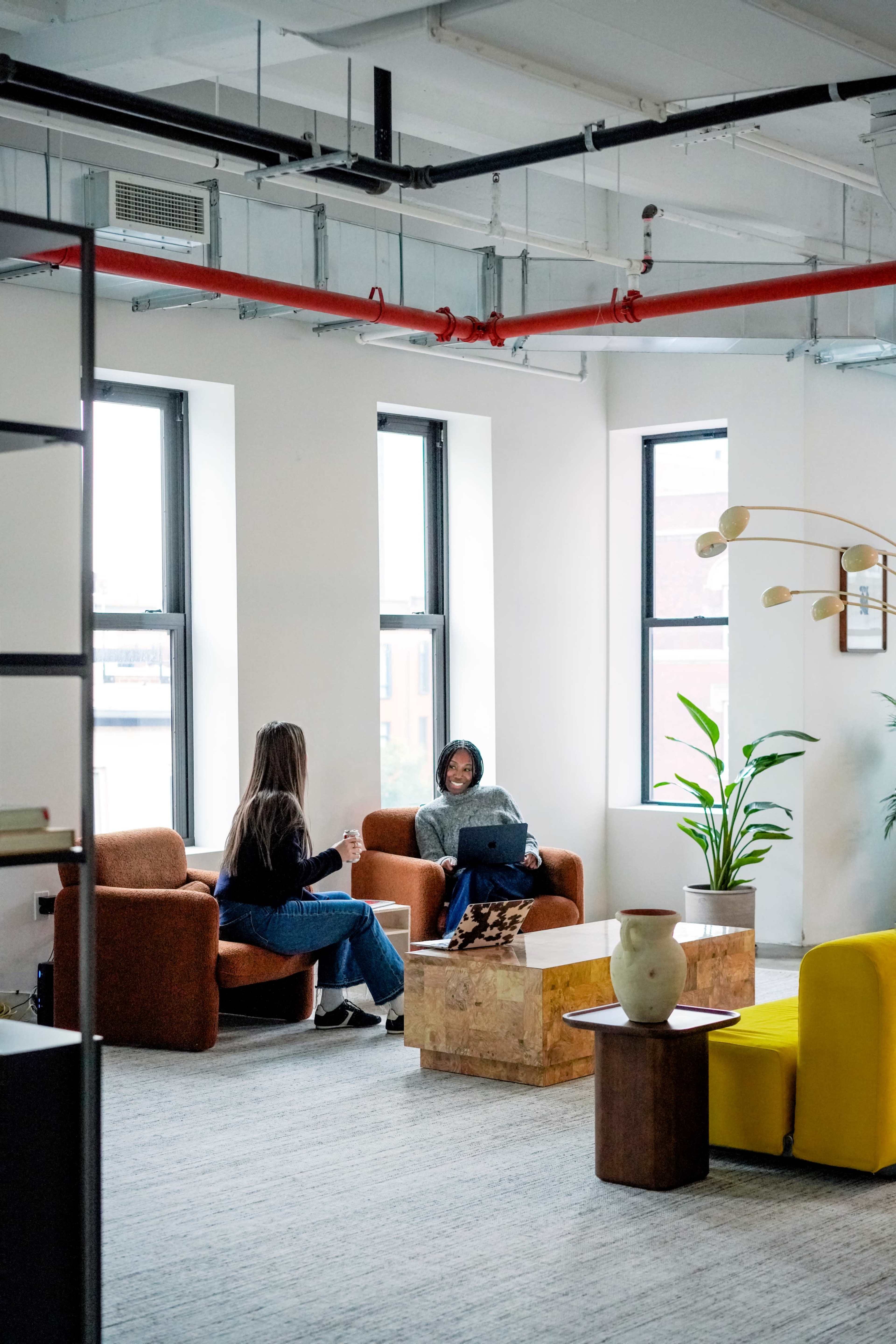 Two people are seated in a well-lit modern office space with large windows, surrounded by plants and minimalist furniture.