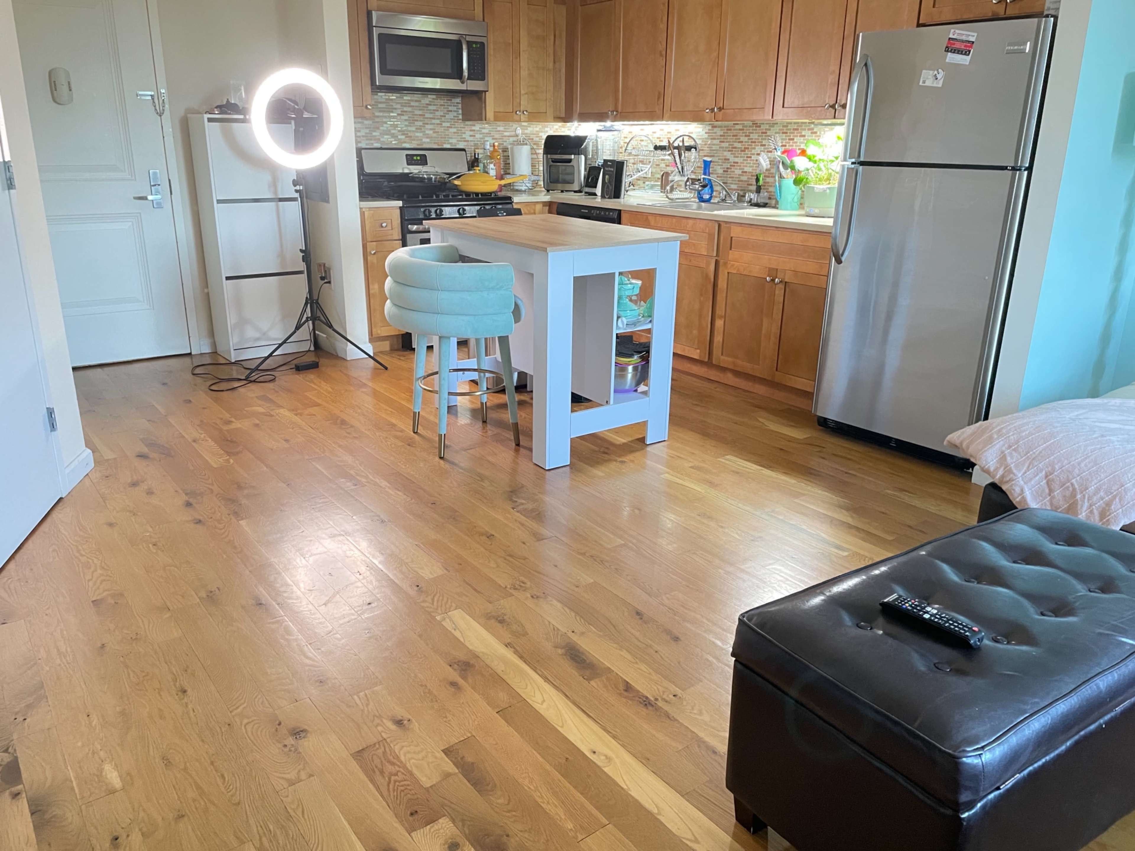 A modern kitchen and living area features wooden floors, light wood cabinets, a stainless steel refrigerator, a ring light, and a small table with stools.