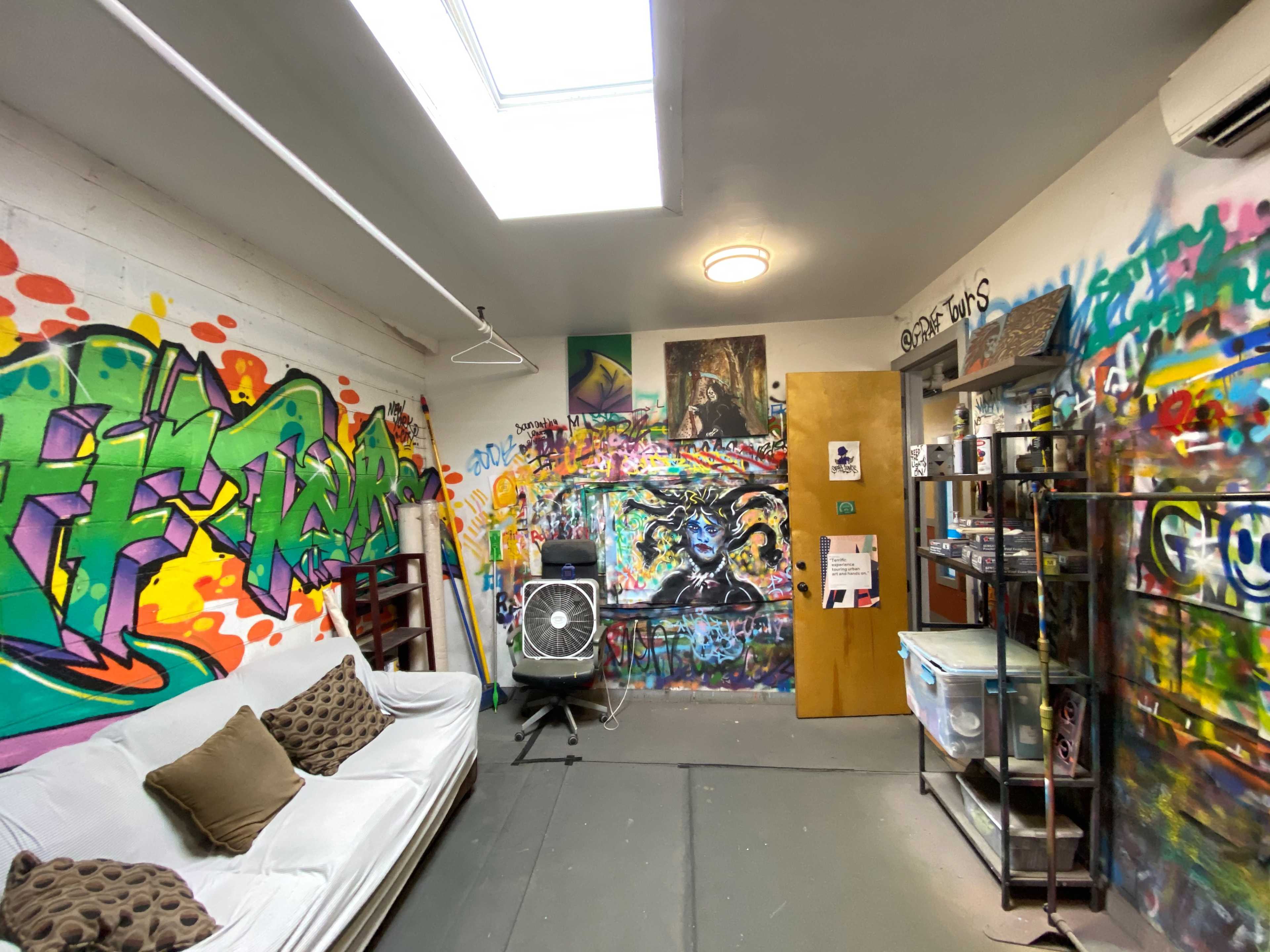 A brightly painted room featuring graffiti on the walls, a white sofa with cushions, and a fan positioned in the corner.