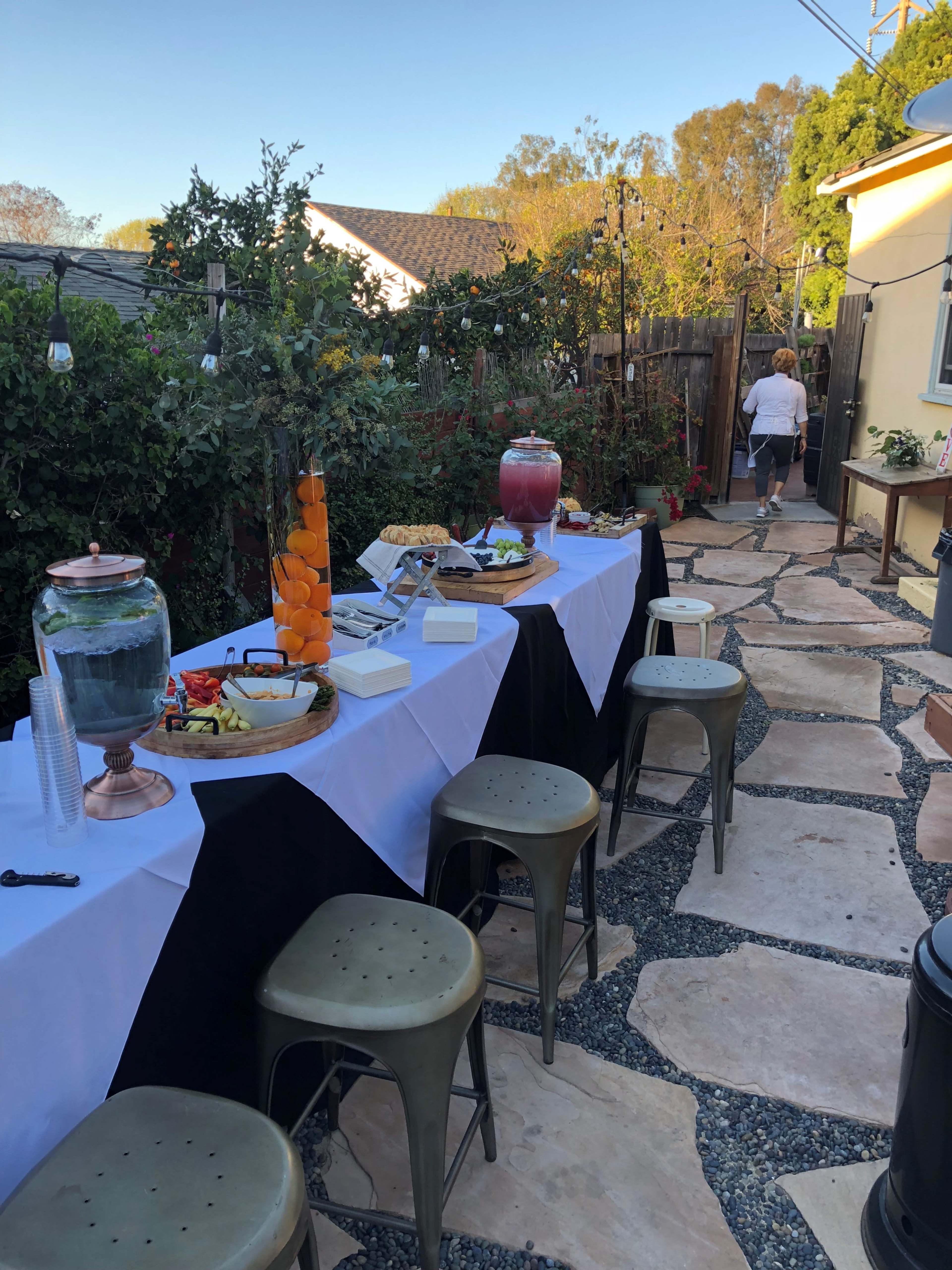 A long table dressed in black and white linens is set with various food items and drinks in a garden, while a person walks along a stone pathway in the background.