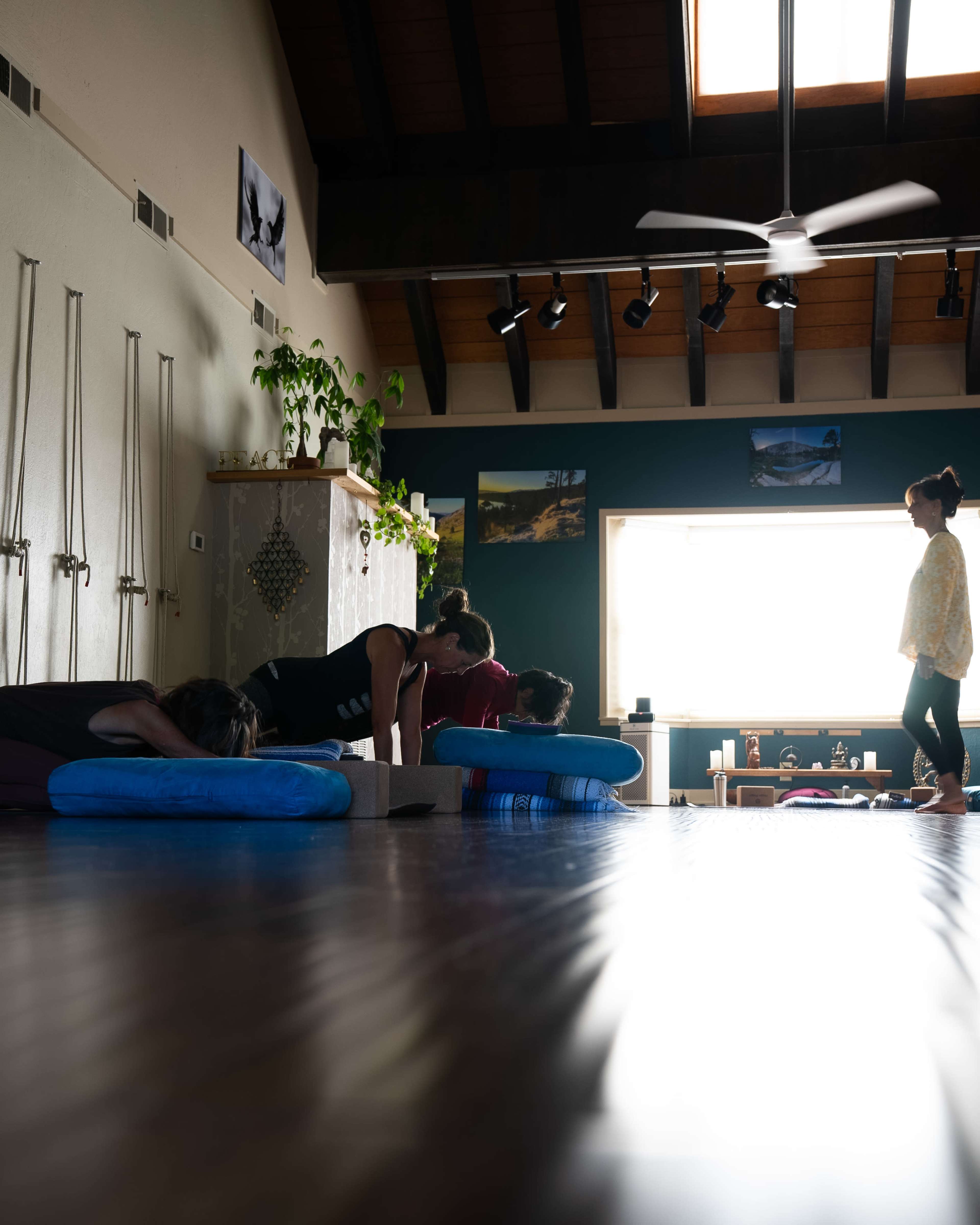 A group of people practice yoga on mats in a spacious room with large windows and wooden beams overhead.