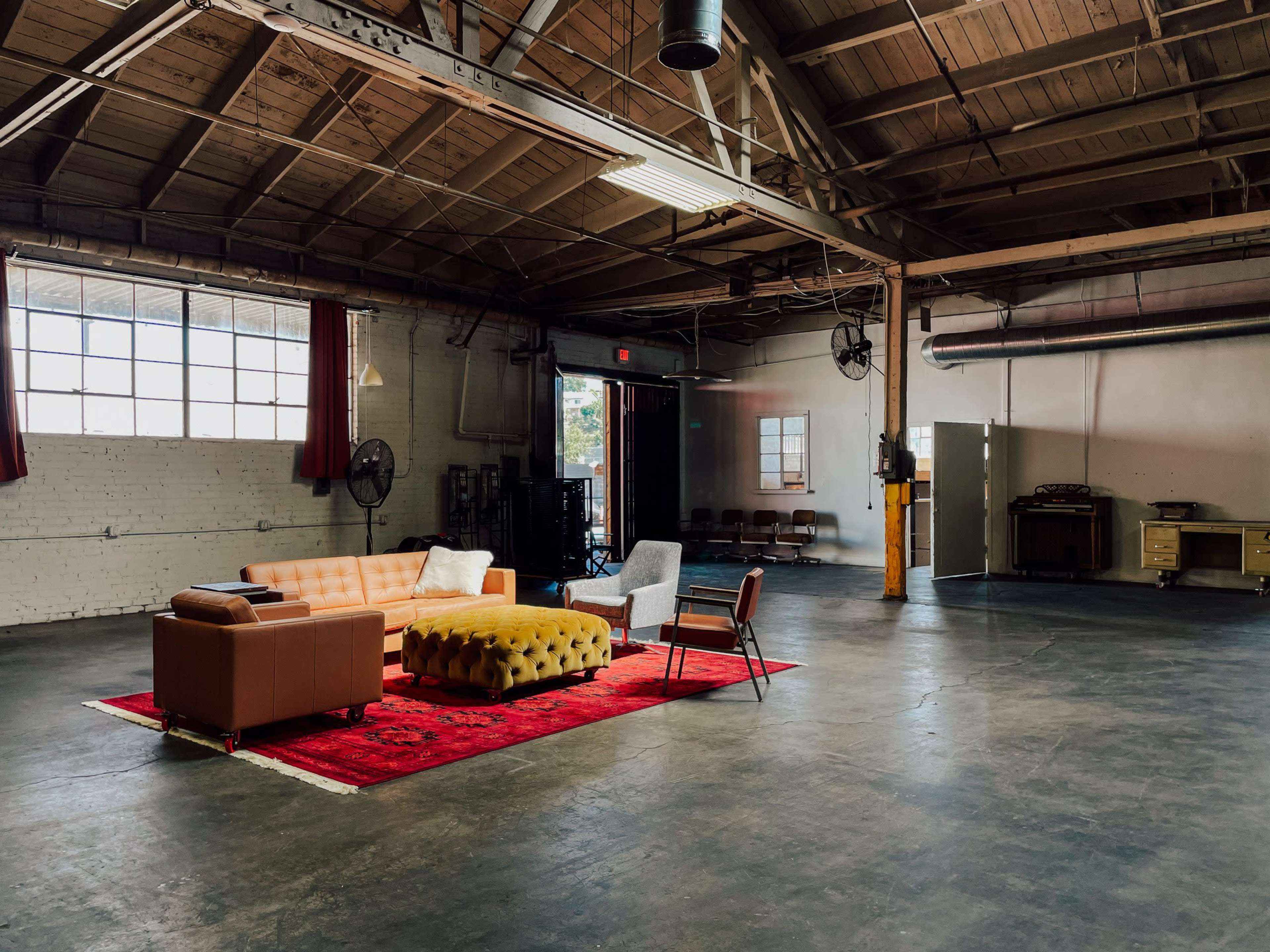 The image shows a spacious industrial-style room featuring a seating area with a couch, an ottoman, and a chair on a large red rug.