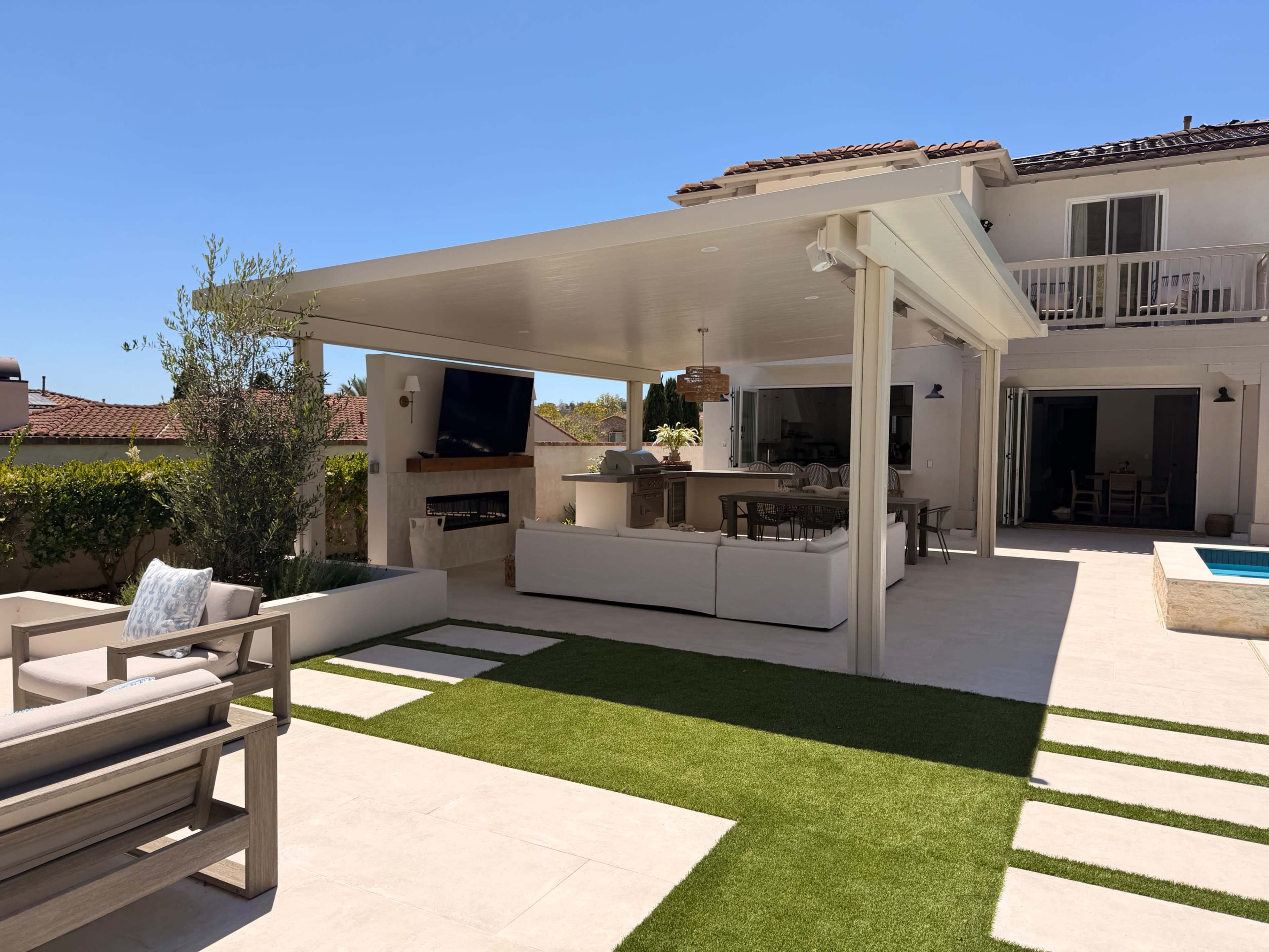 The image shows a patio area of a modern home featuring a large covered seating space with outdoor furniture and a swimming pool in the background.