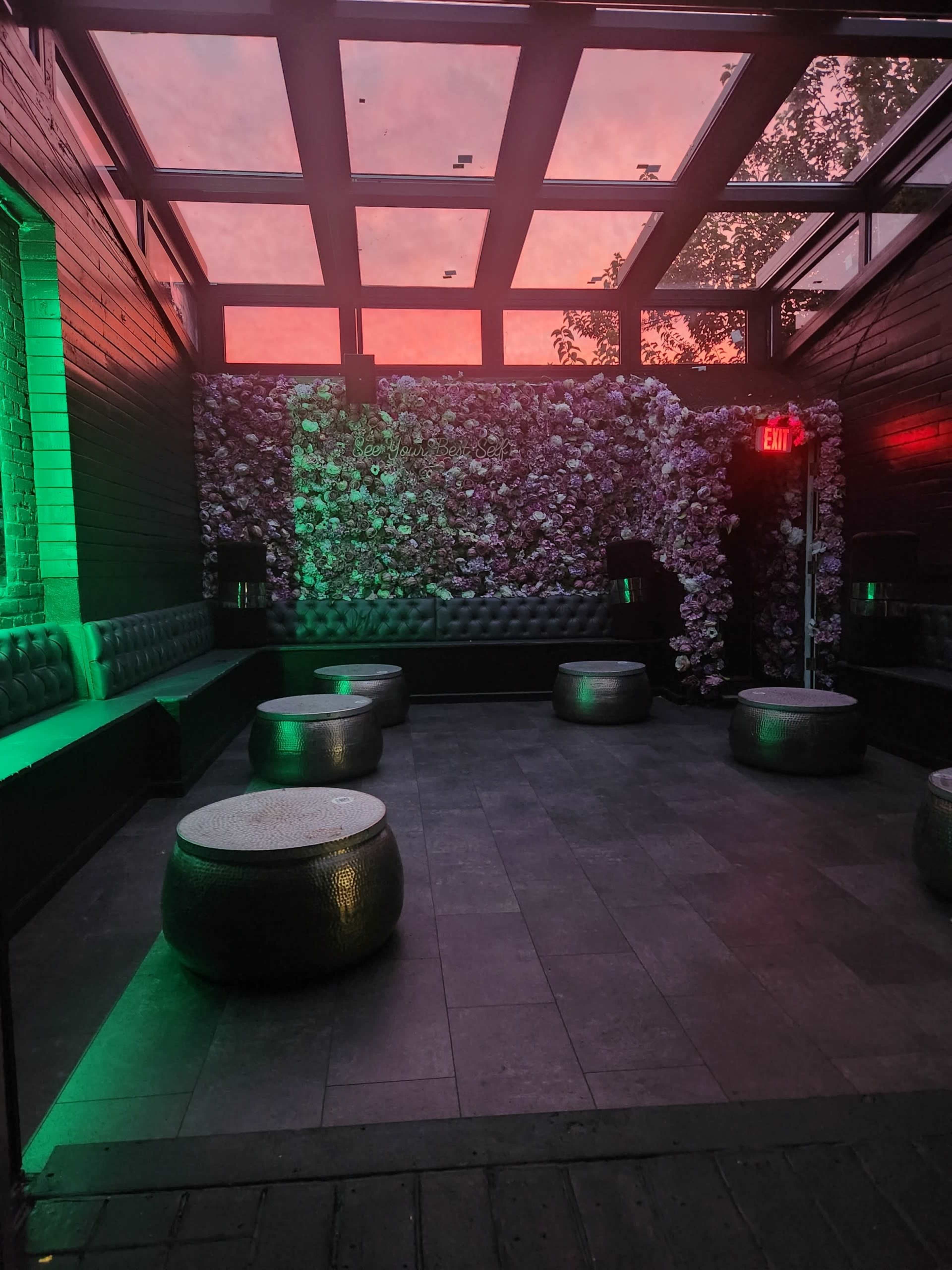 An empty lounge area with floral wall decorations, low seating, and illuminated green lights under a pink-hued sky.