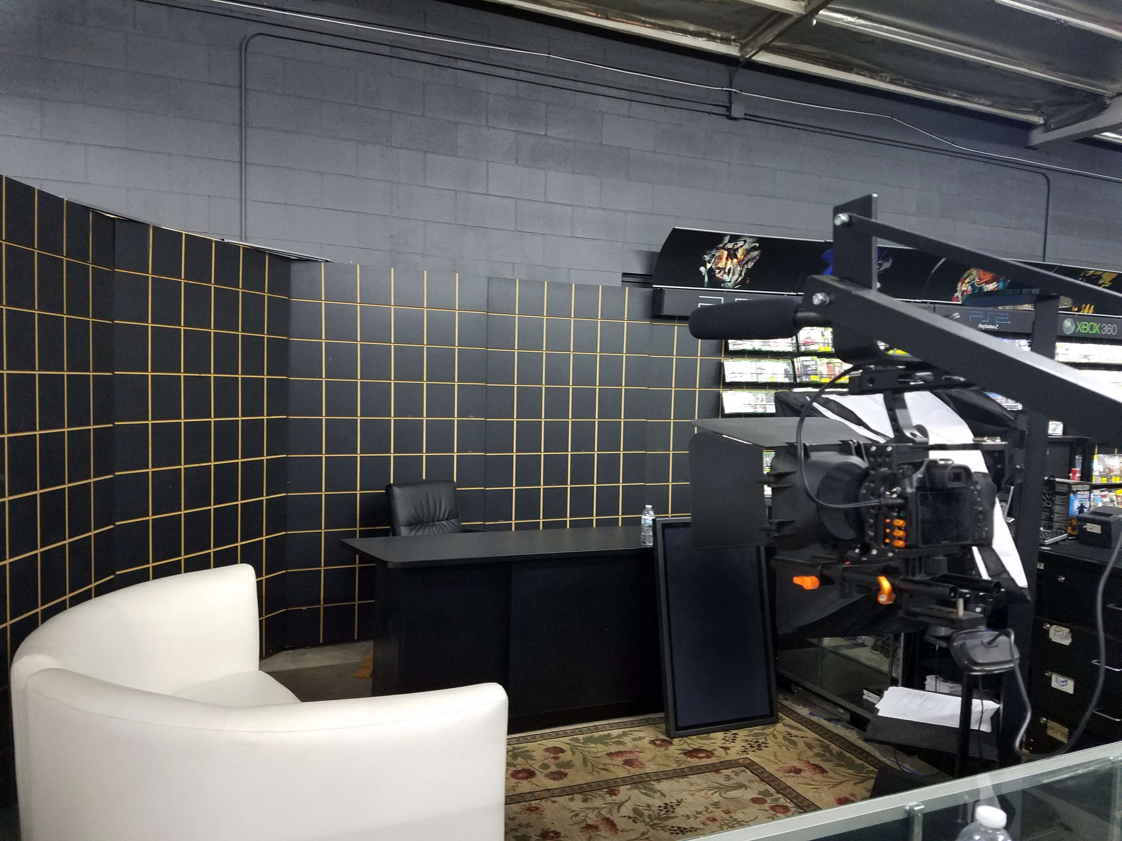 A video production setup featuring a white couch, a desk, and a camera on a tripod against a backdrop of black and orange grid patterns.