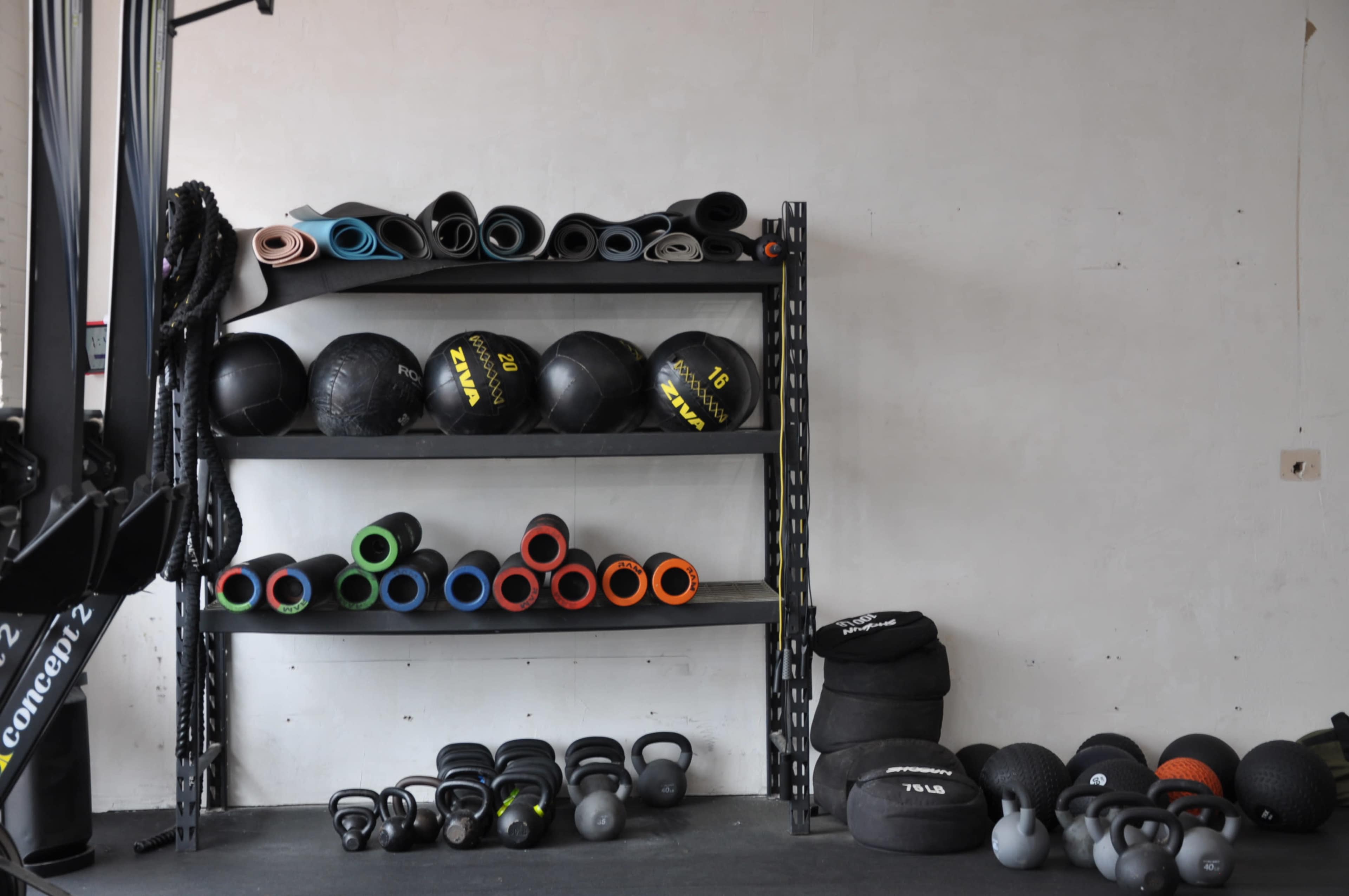 A workout area with two metal shelves stacked with various exercise balls and foam rollers, alongside a collection of kettlebells and weights on the floor.