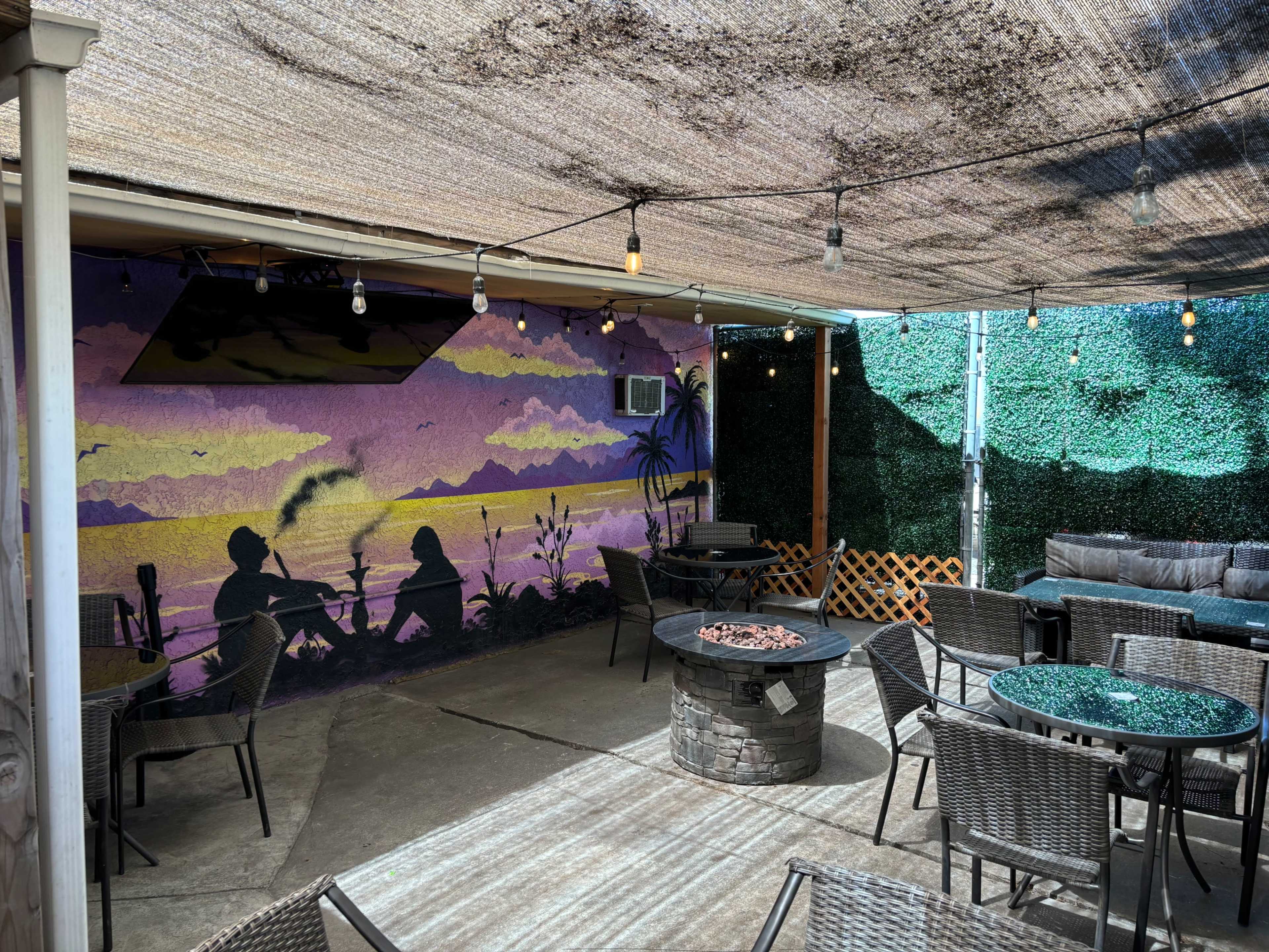A patio area with a mural, outdoor seating, and a fire pit, all under a shaded canopy.