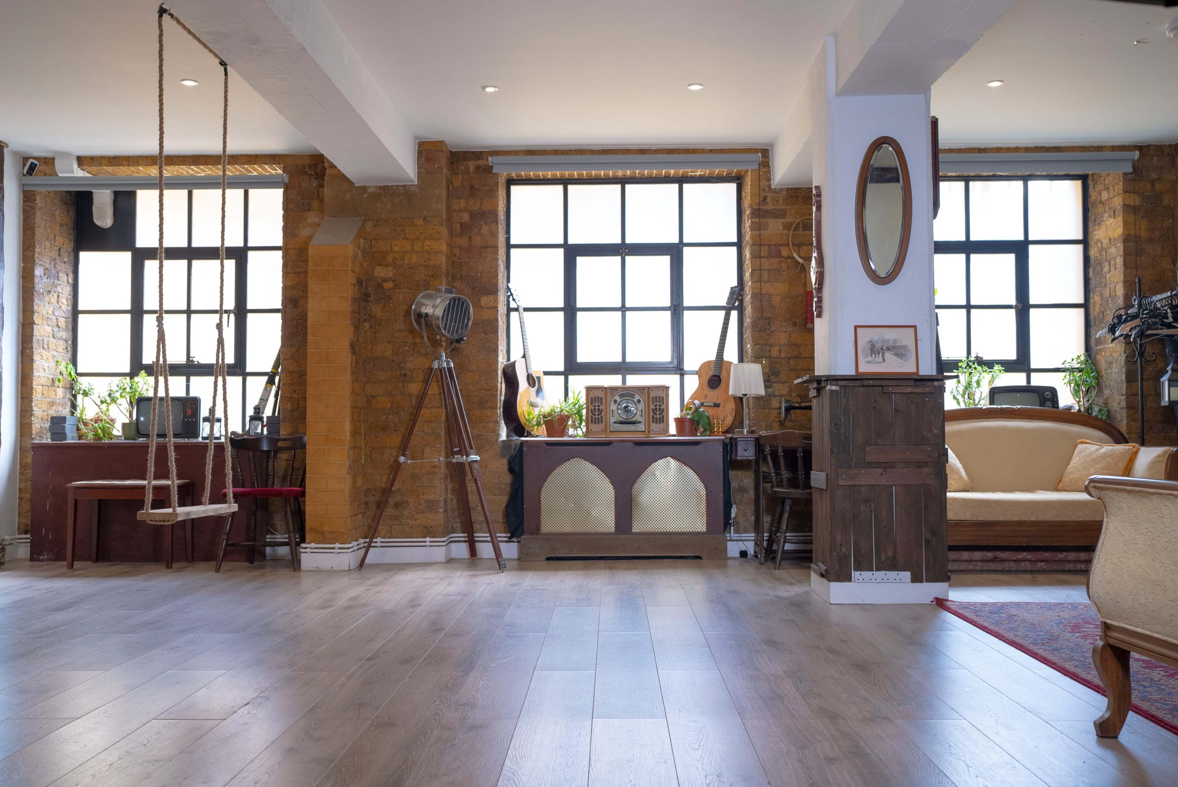 A spacious interior with wooden flooring, large windows, and vintage decor, including a swing, guitars, and plants.