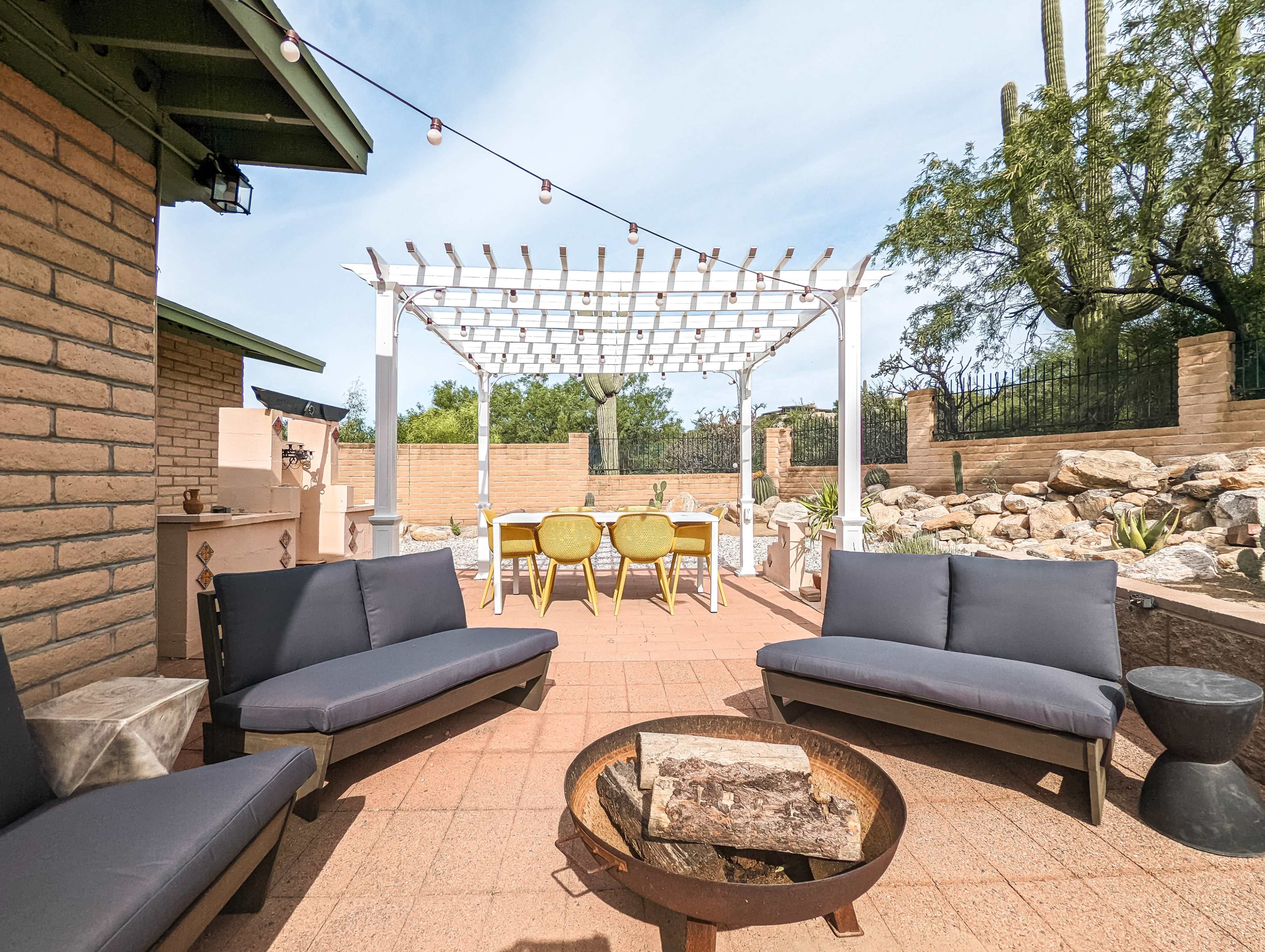 A patio area features grey outdoor sofas surrounding a fire pit, with a pergola overhead and a set of dining chairs at a table in the background.