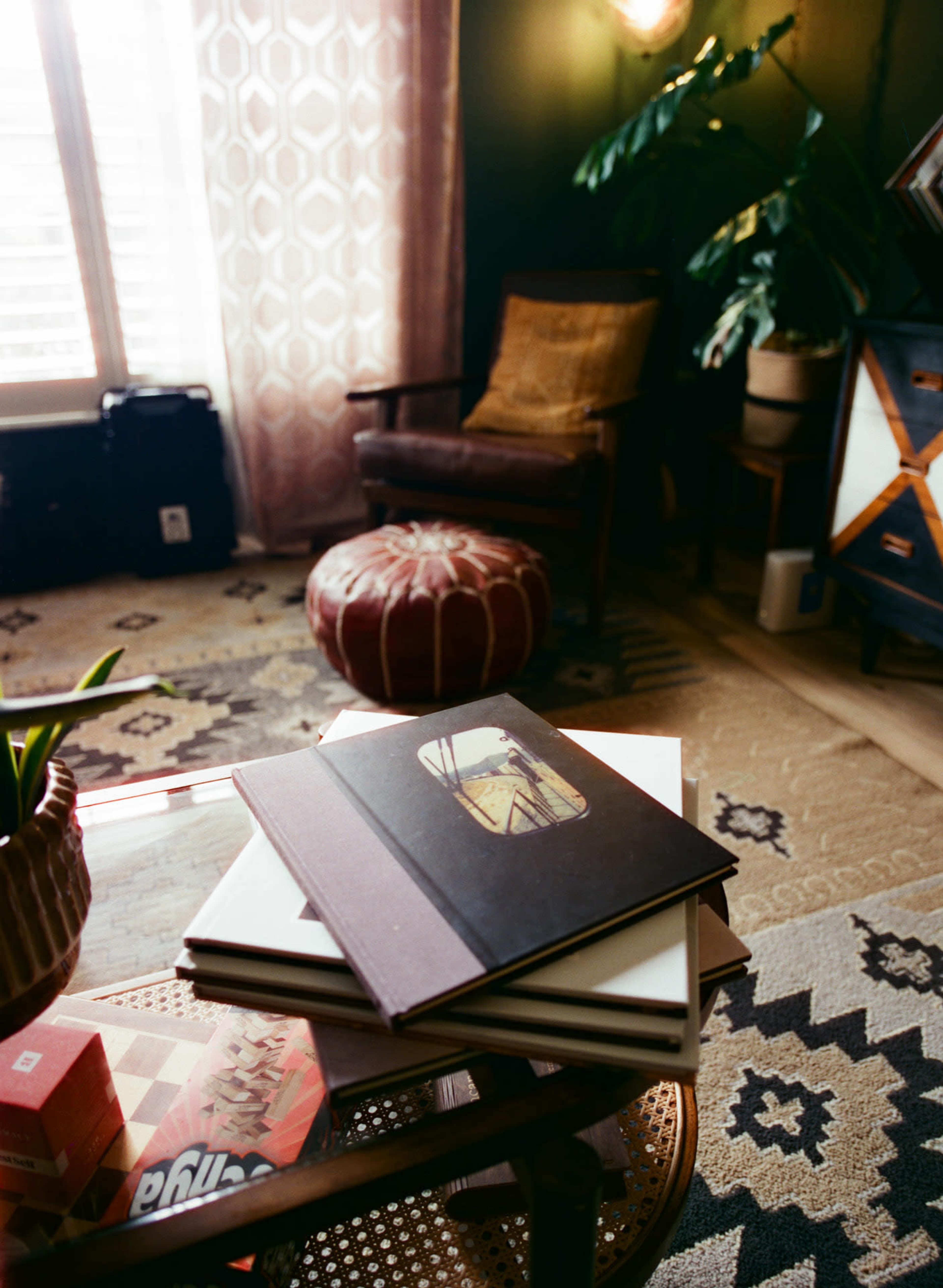 The image shows a cozy corner of a room with a stack of photo albums on a glass coffee table, a patterned rug, an armchair, and a potted plant near a window.