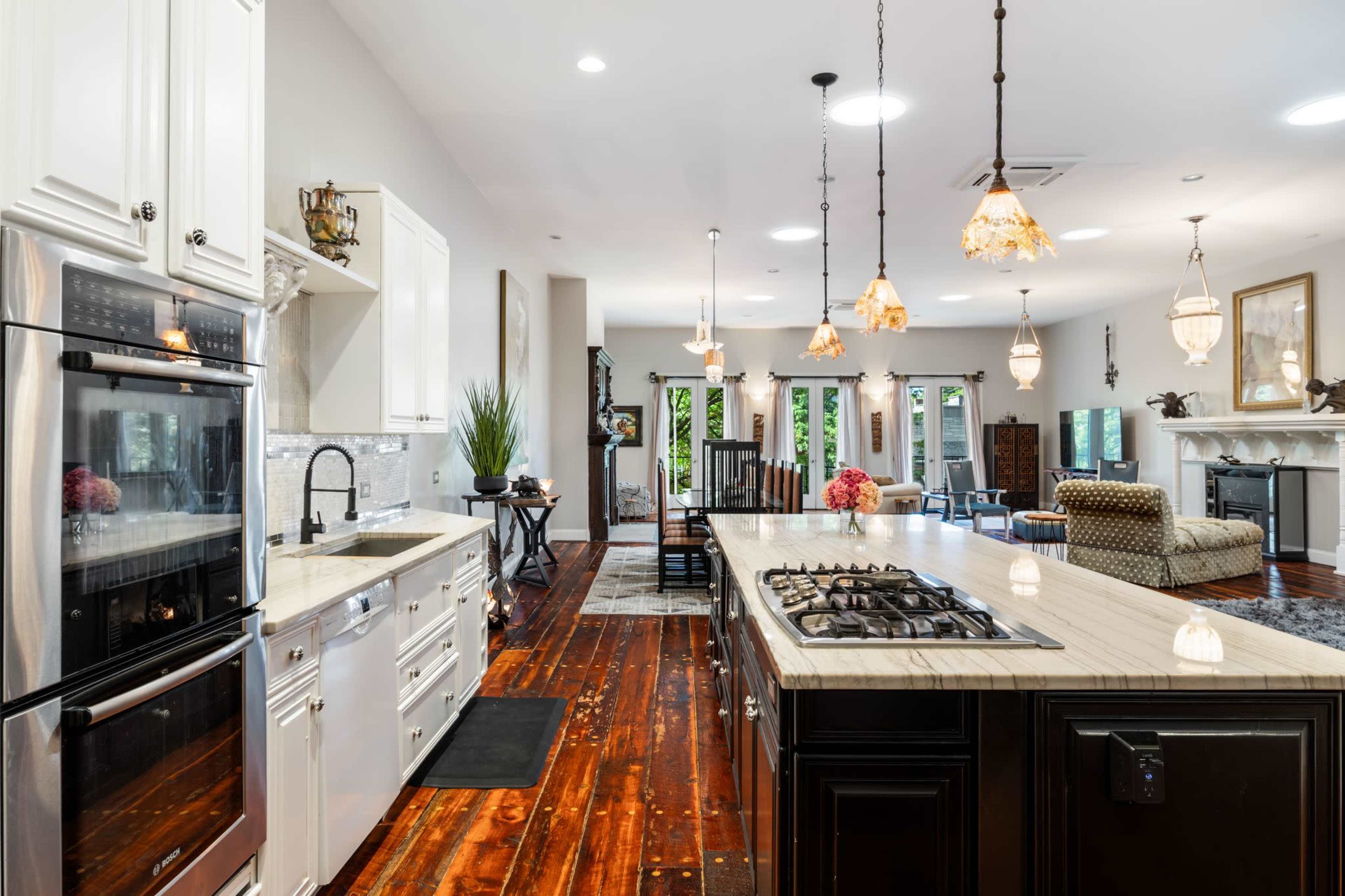 The image shows a spacious kitchen with an island, featuring a combination of white and black cabinetry, a gas stove, and hardwood floors, leading into a living area with comfortable seating and natural light.