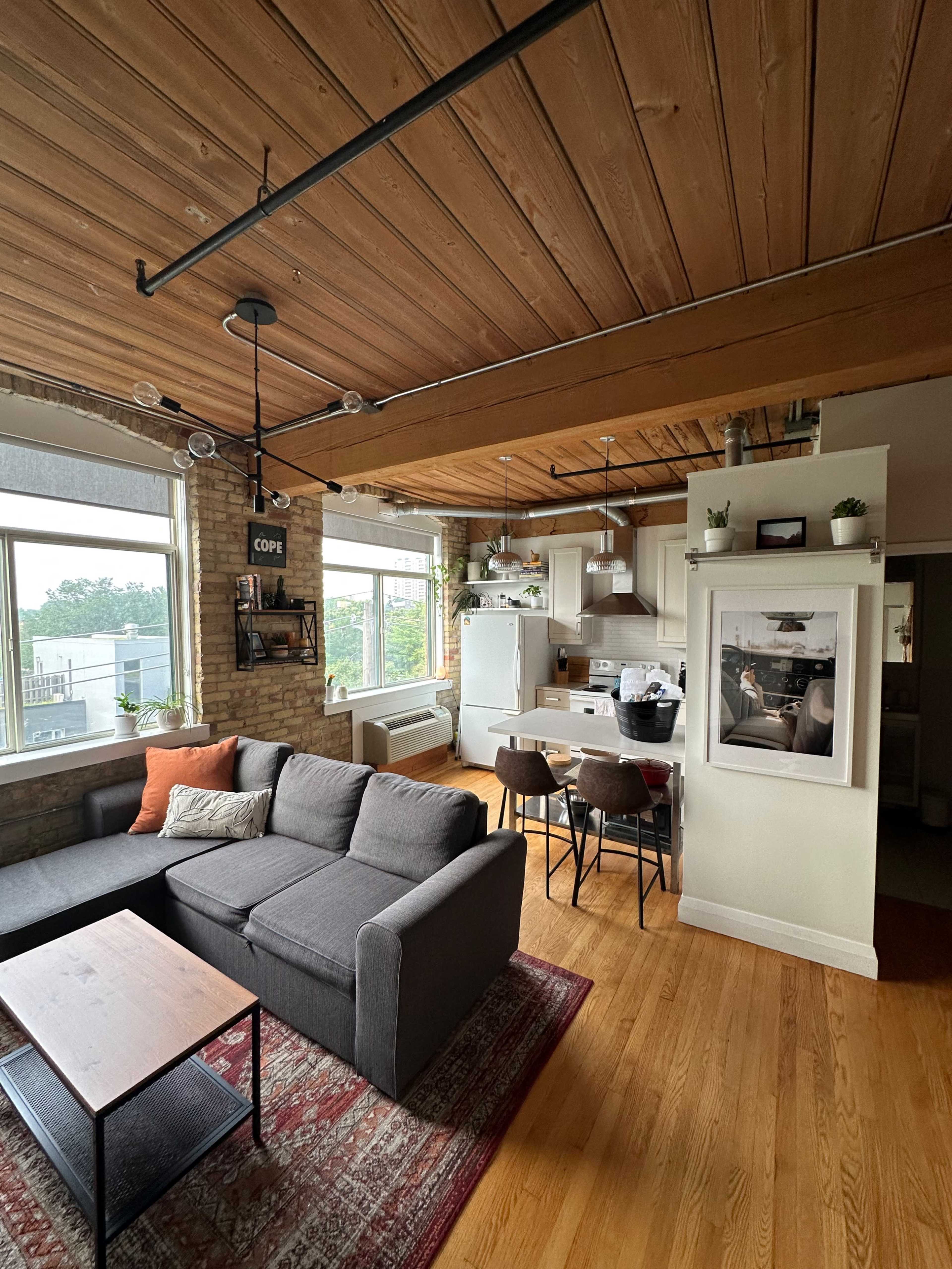 A modern, open-plan living space featuring a gray sofa, a wooden dining table, and large windows with natural light.
