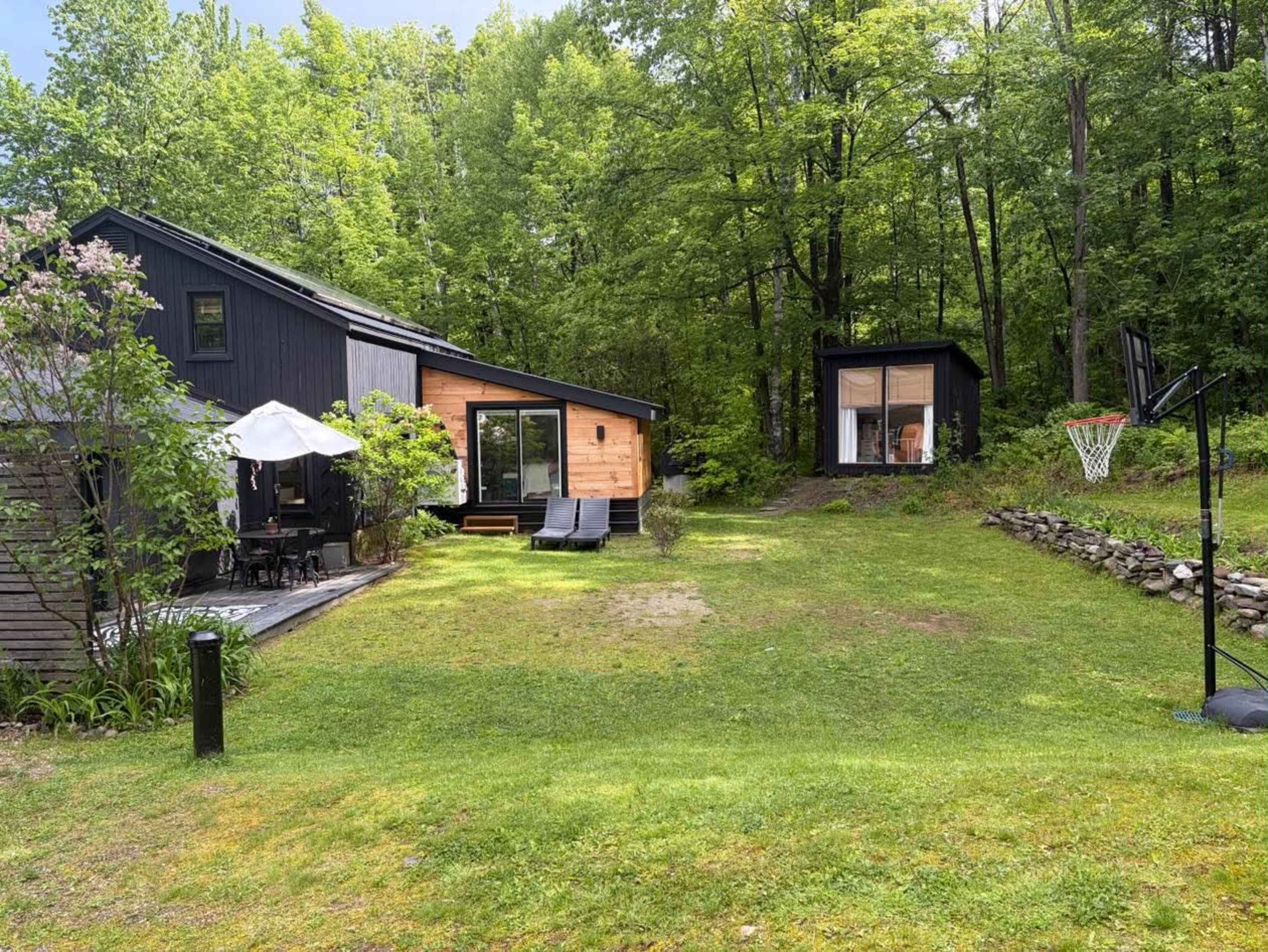 A modern house with a wooden facade and a separate small structure is set in a lush, green landscape surrounded by trees.