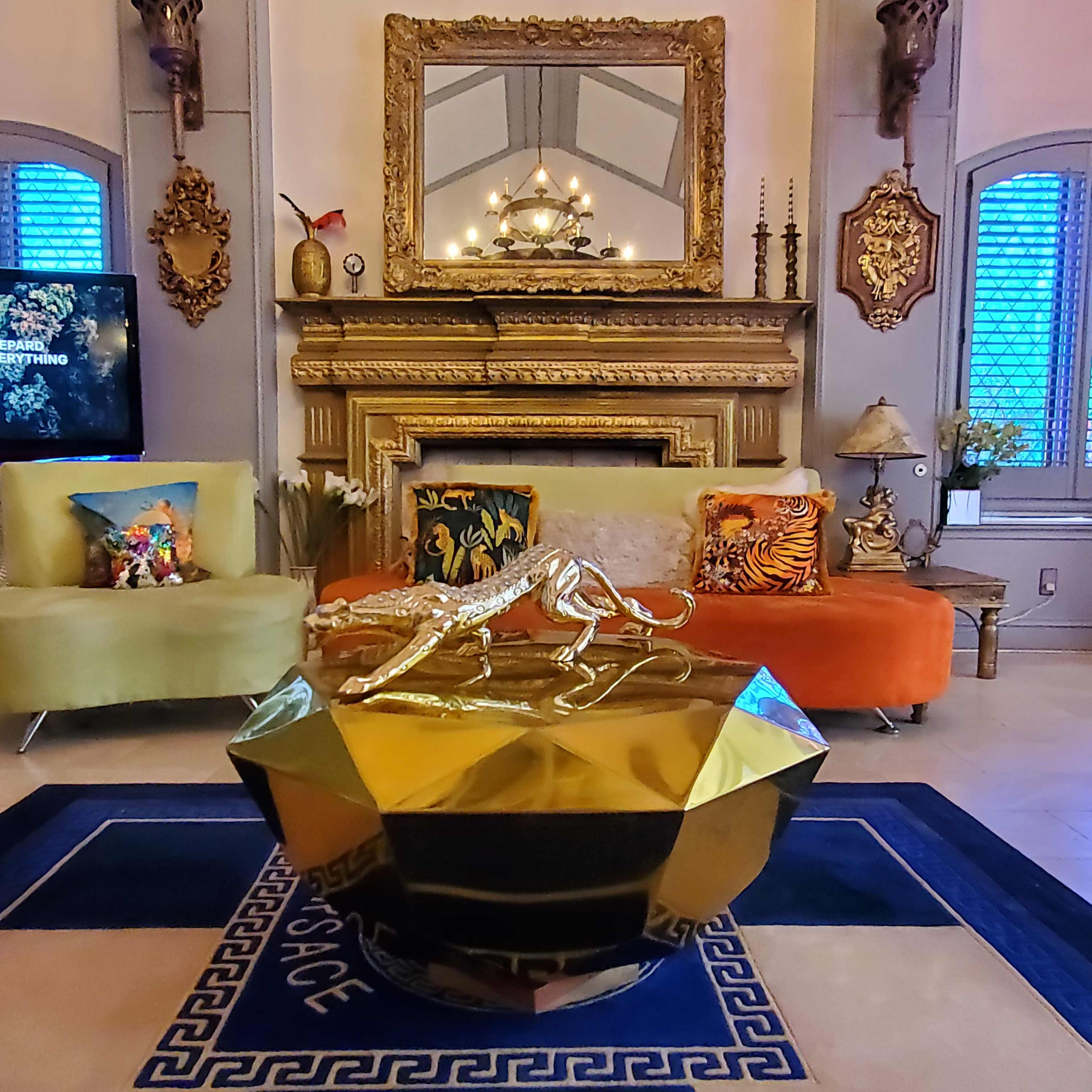The image shows a luxurious living room with a golden table featuring a leopard sculpture, colorful sofas, and ornate decor surrounding a fireplace.