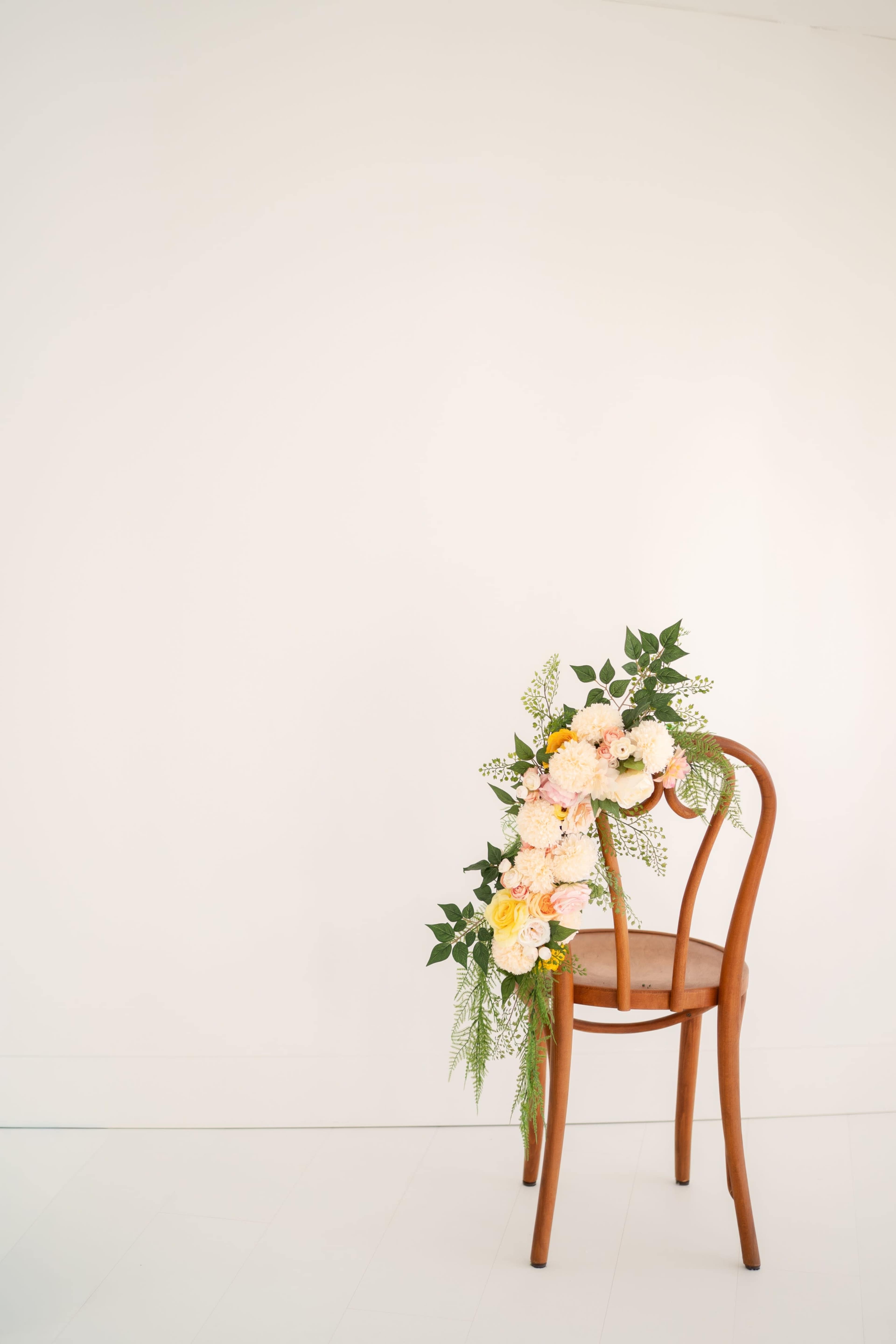 A wooden chair is adorned with a floral arrangement of pink and white flowers cascading down its side against a plain white wall.