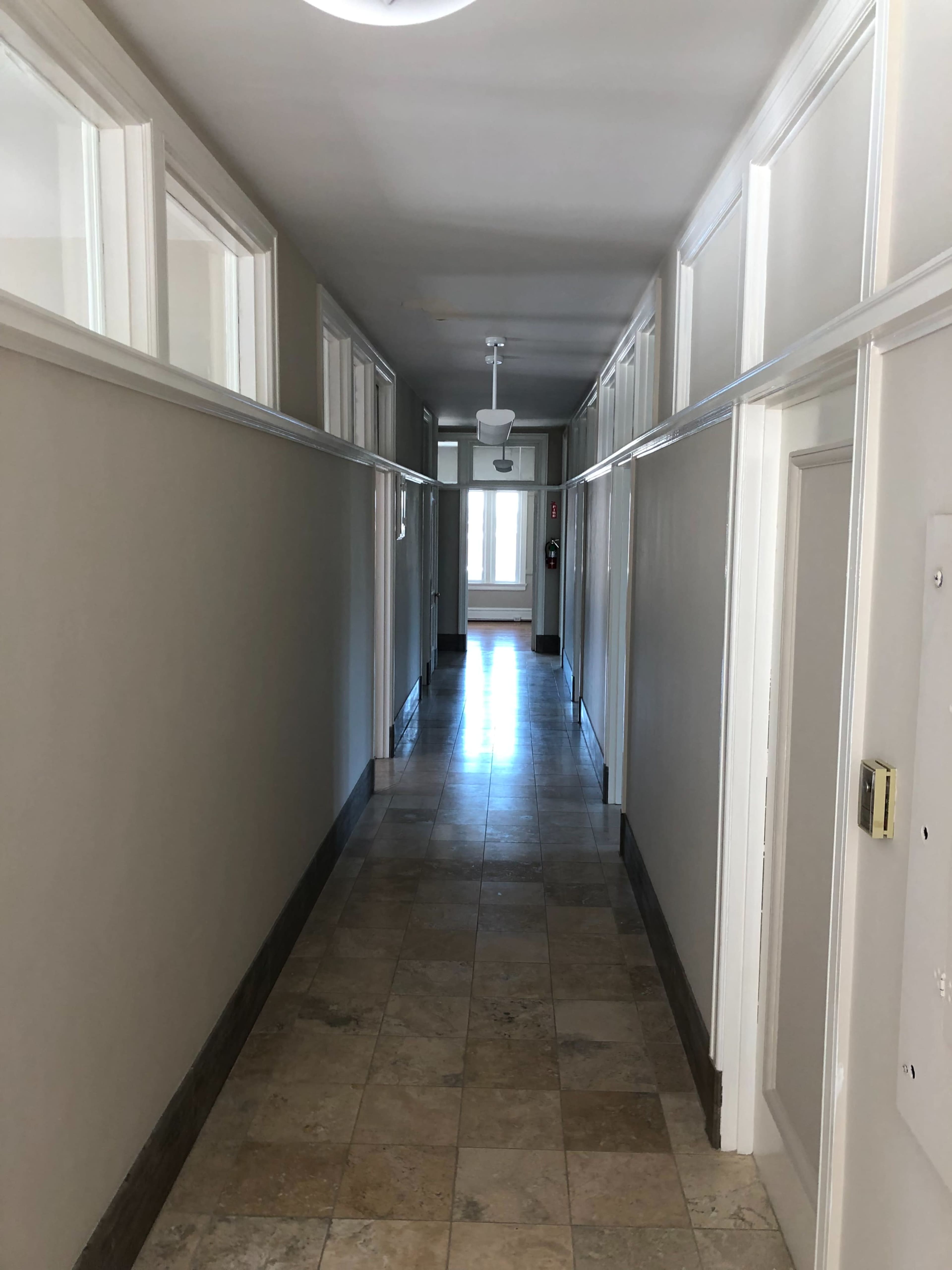 A long, narrow hallway with tile flooring and doors along both sides, leading to a bright area at the end.