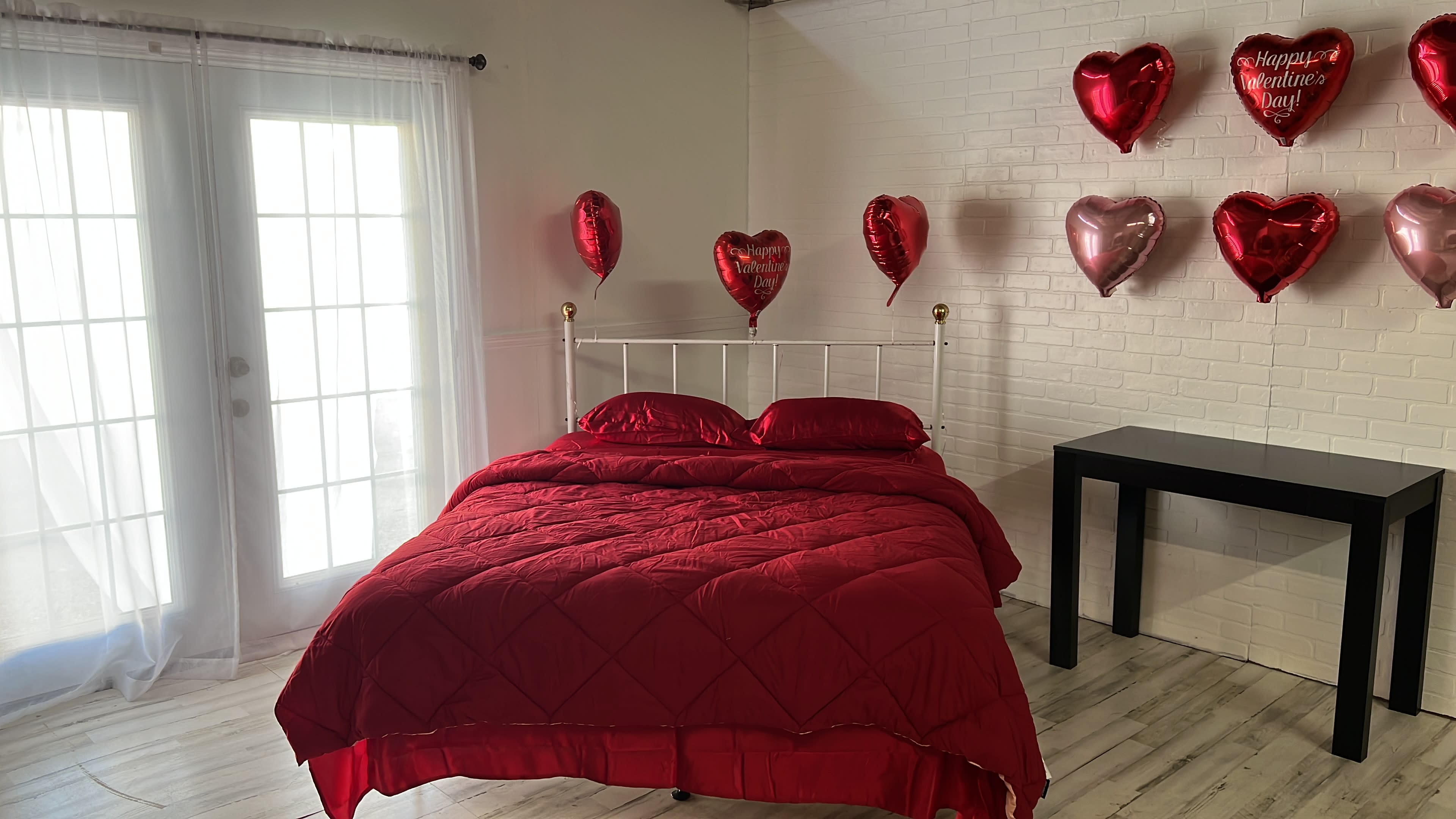 A bedroom features a red bedspread on a white metal bed, adorned with heart-shaped balloons hanging on the wall and a small black table beside it.