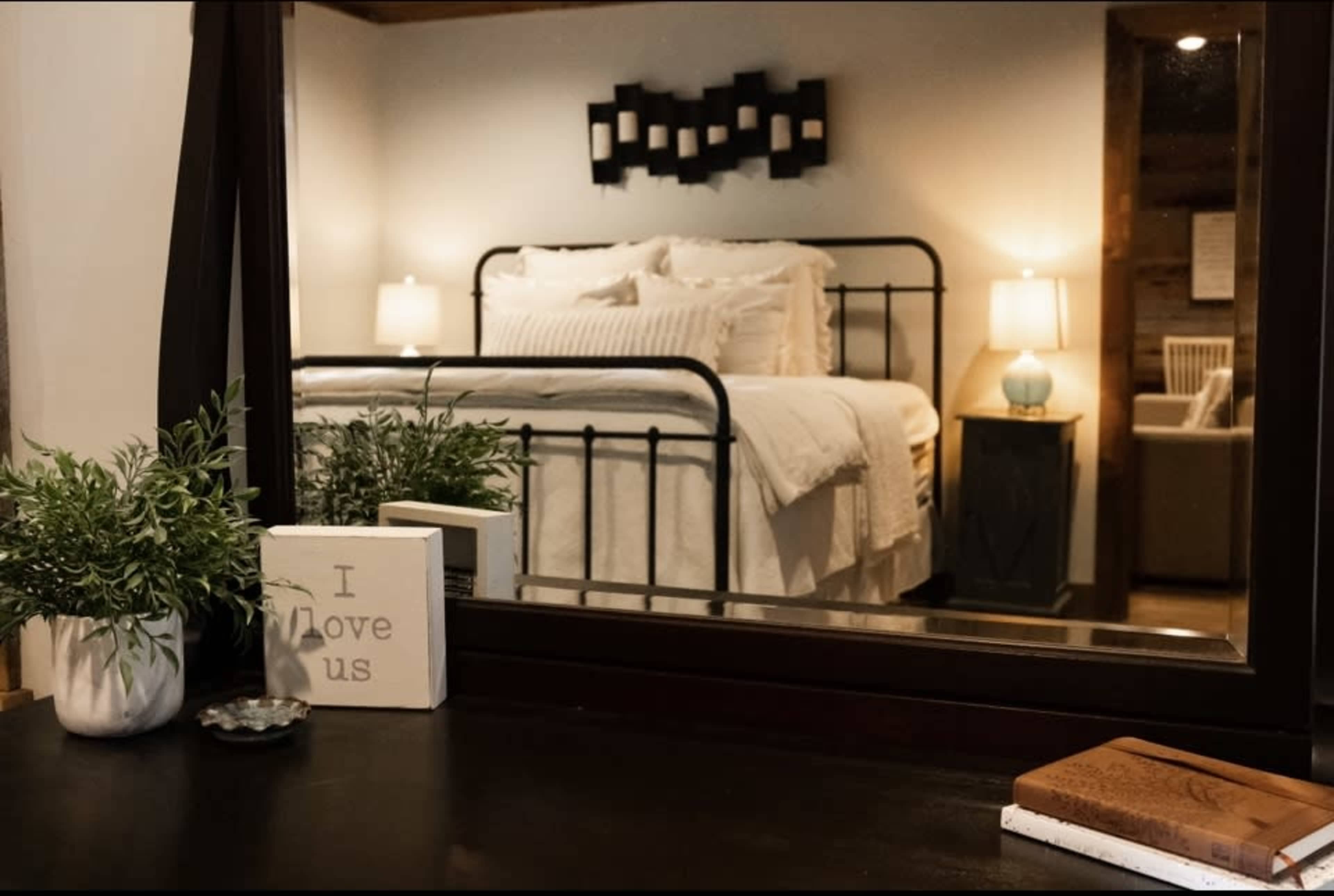 A cozy bedroom with a black metal bed frame, decorative wall art, and bedside lamps, reflected in a dark wooden mirror.