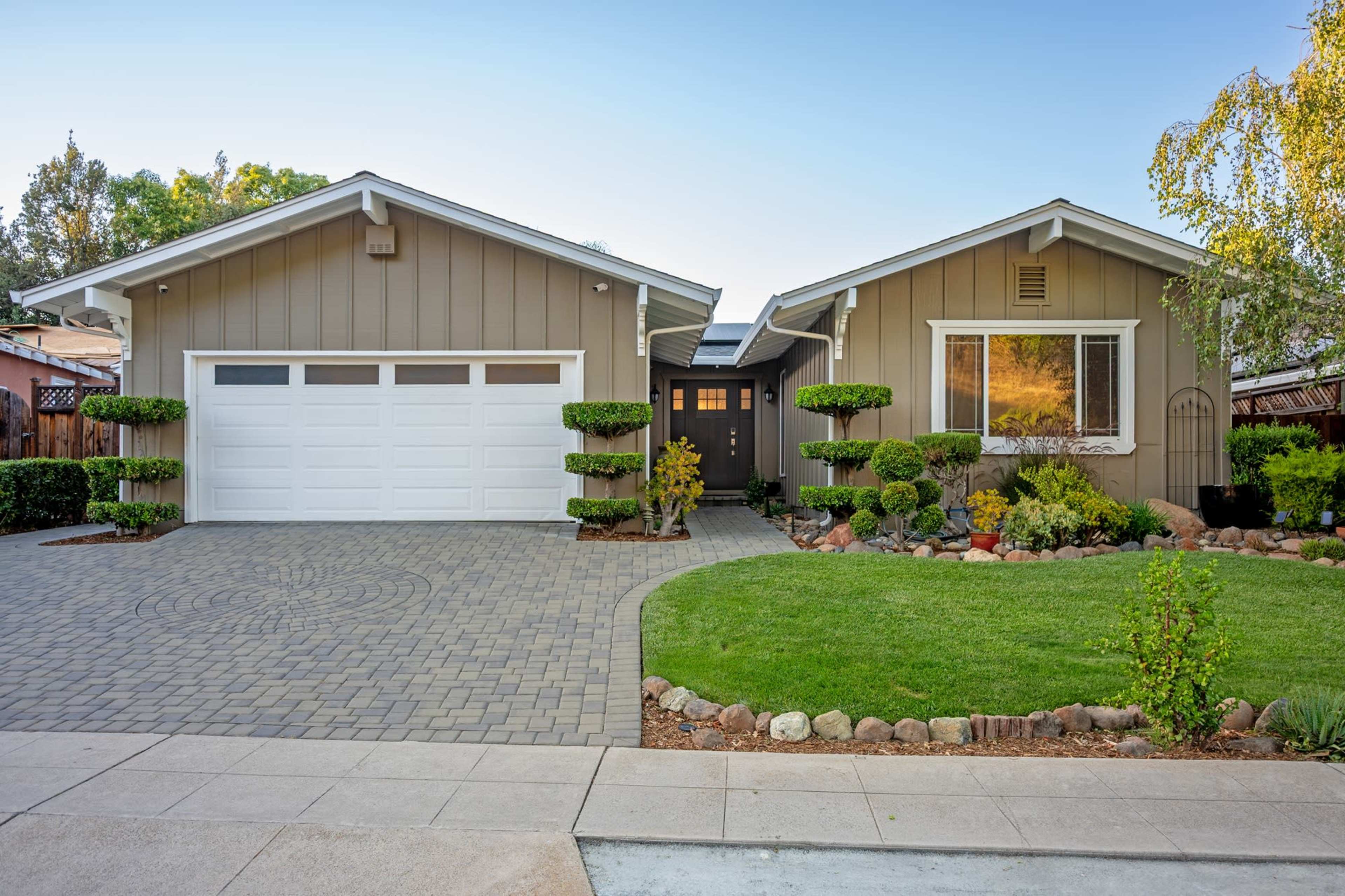 The image shows a modern single-story house with a well-manicured lawn, a paved driveway, and neatly trimmed hedges on either side of the entrance.