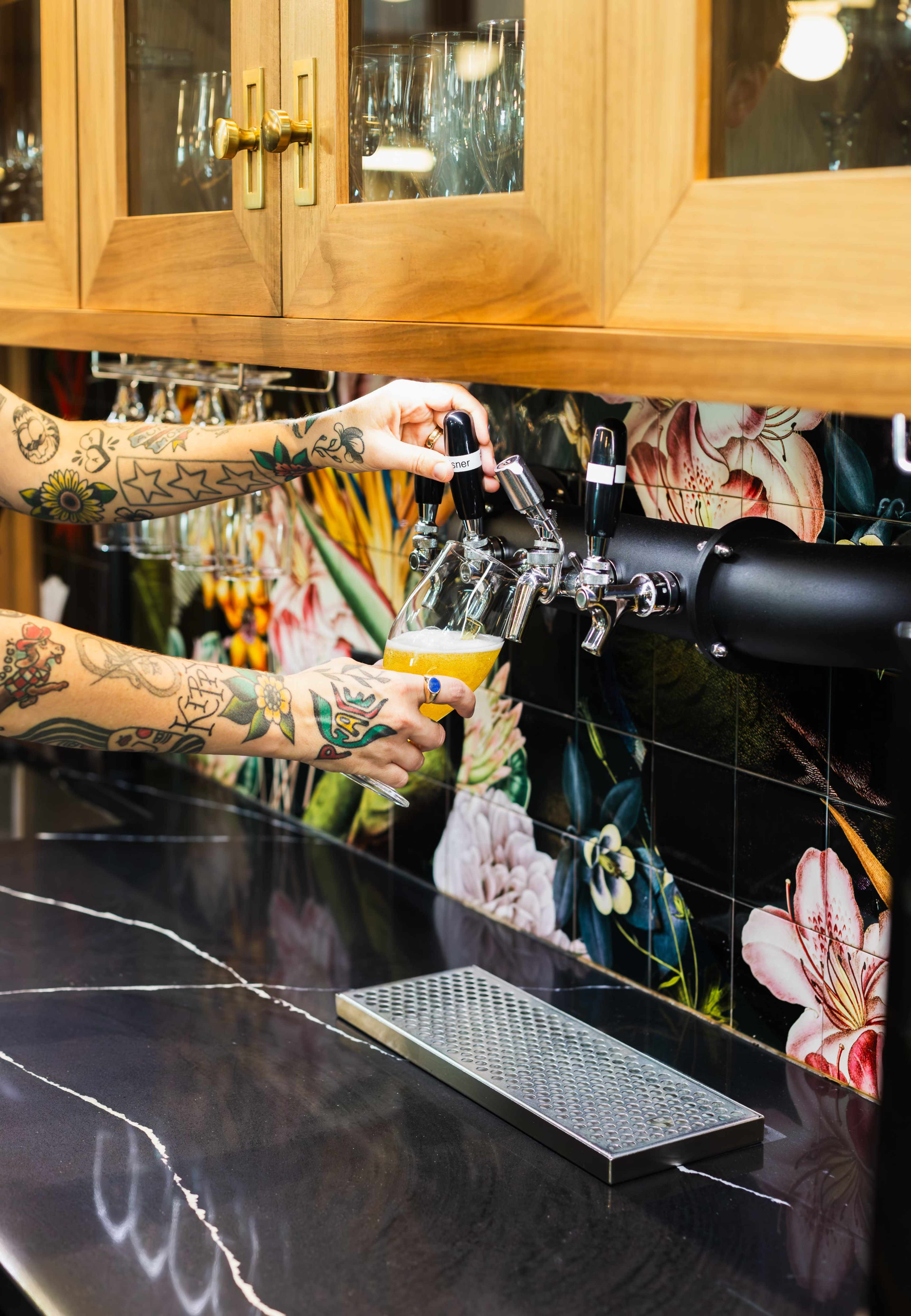 A person with tattoos pours a drink from a tap into a glass at a bar with floral-themed tiles and wooden cabinetry.
