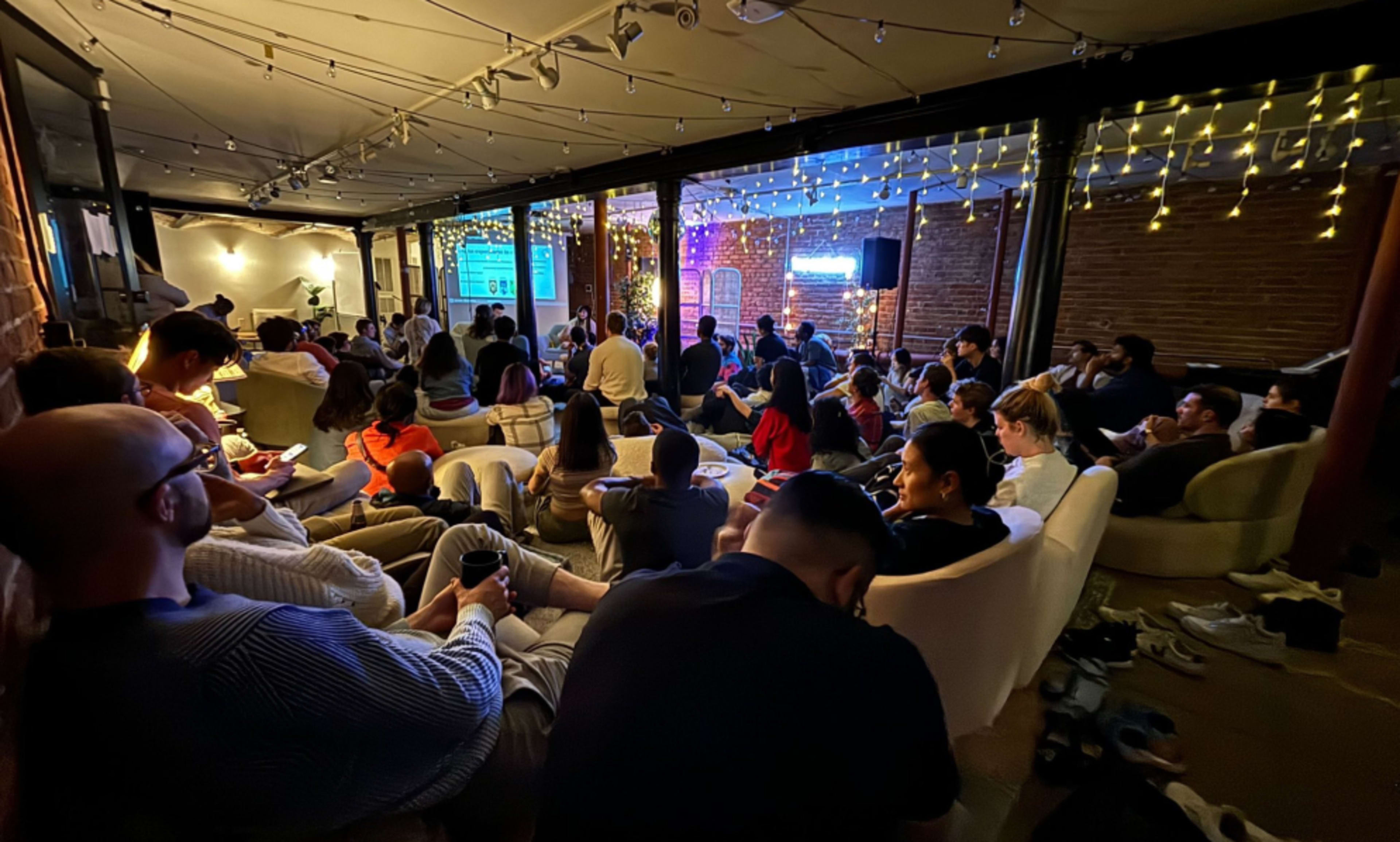 A diverse group of people is seated in a cozy, brightly lit space, engaged in a presentation displayed on a screen at the front.