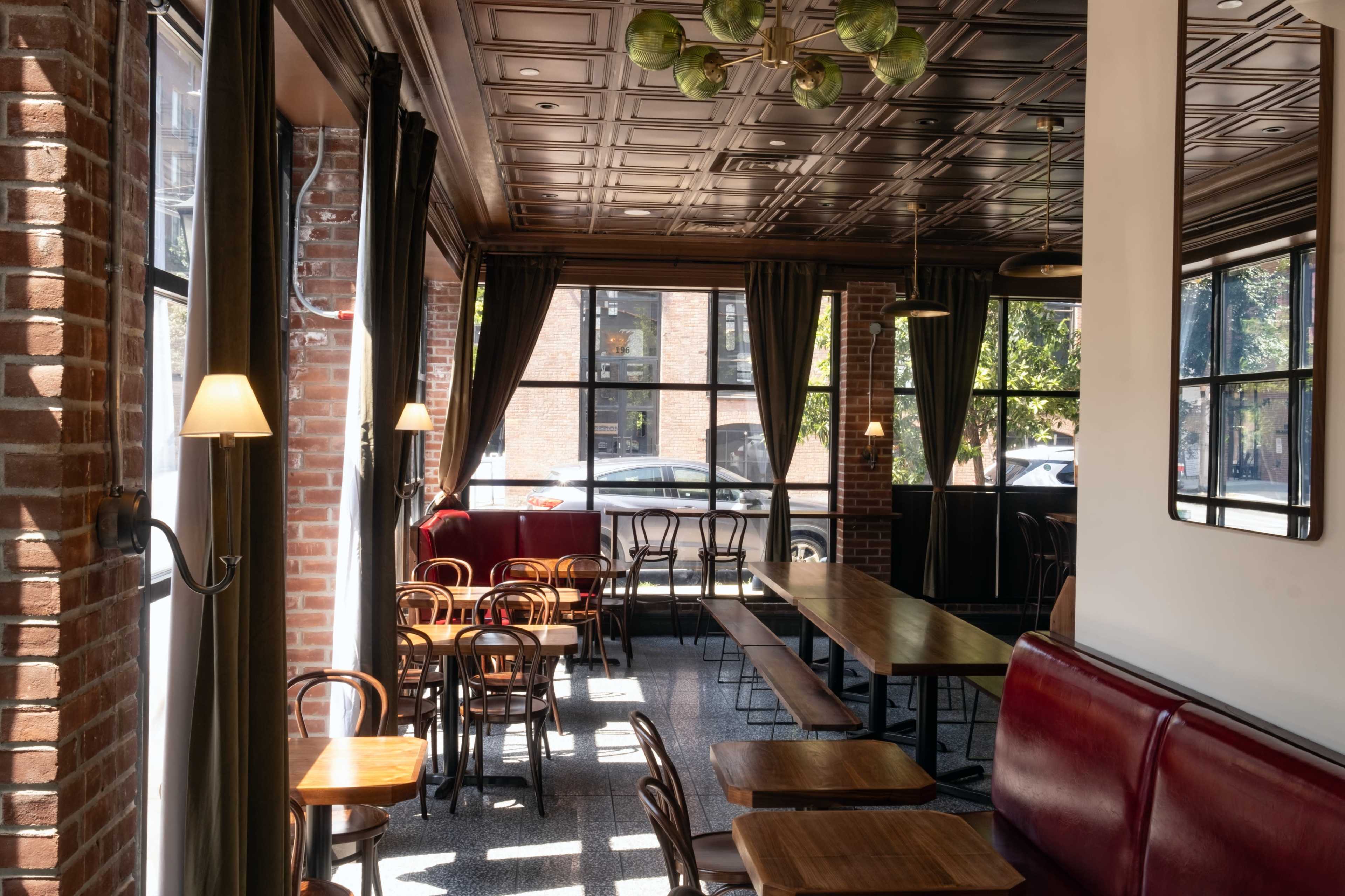 The image shows a well-lit café interior with wooden tables and chairs, large windows, and brick walls.