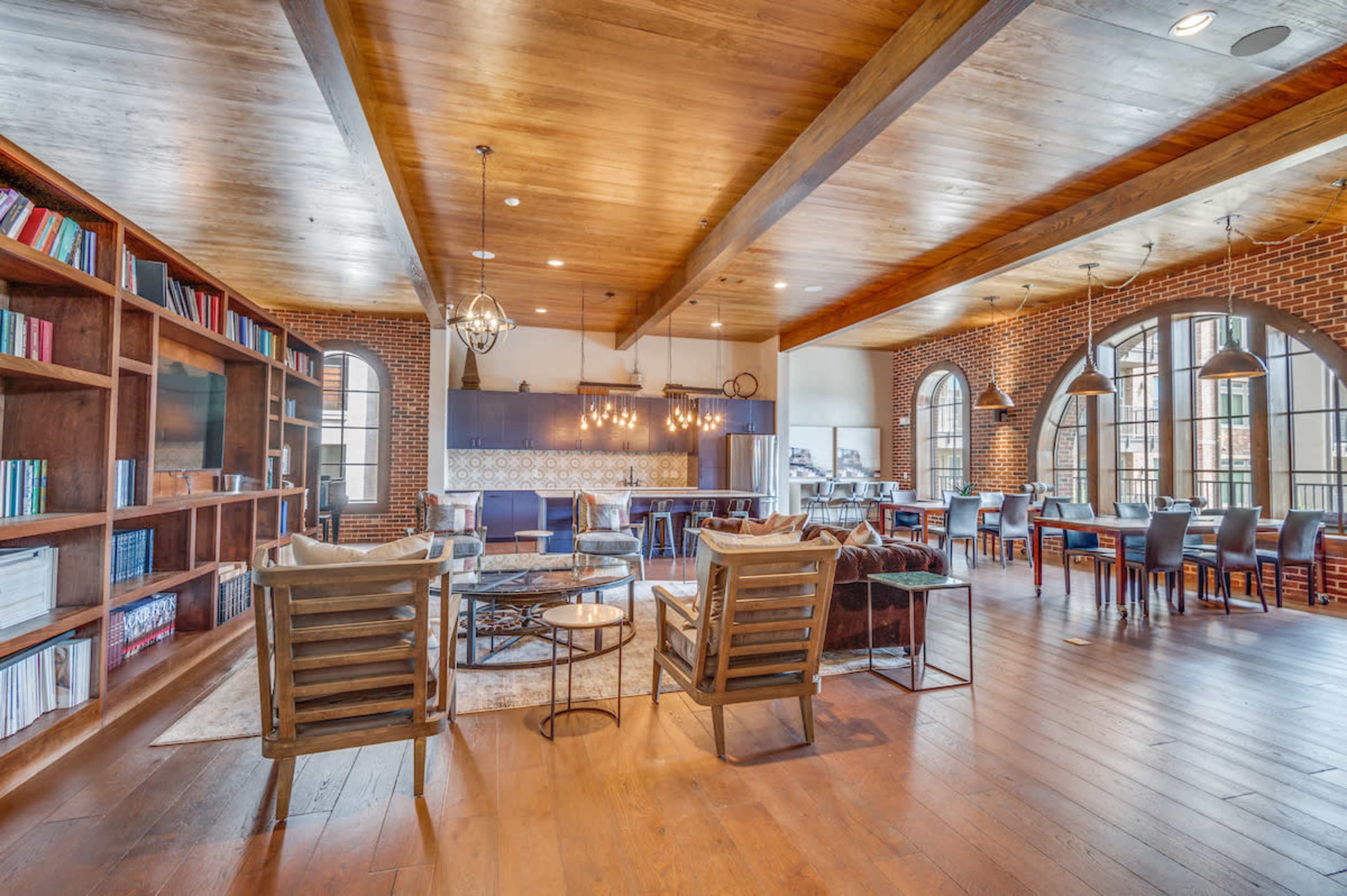 A spacious, modern lounge with wooden beams, a large bookshelf, and a dining area featuring arched windows and a kitchen in the background.