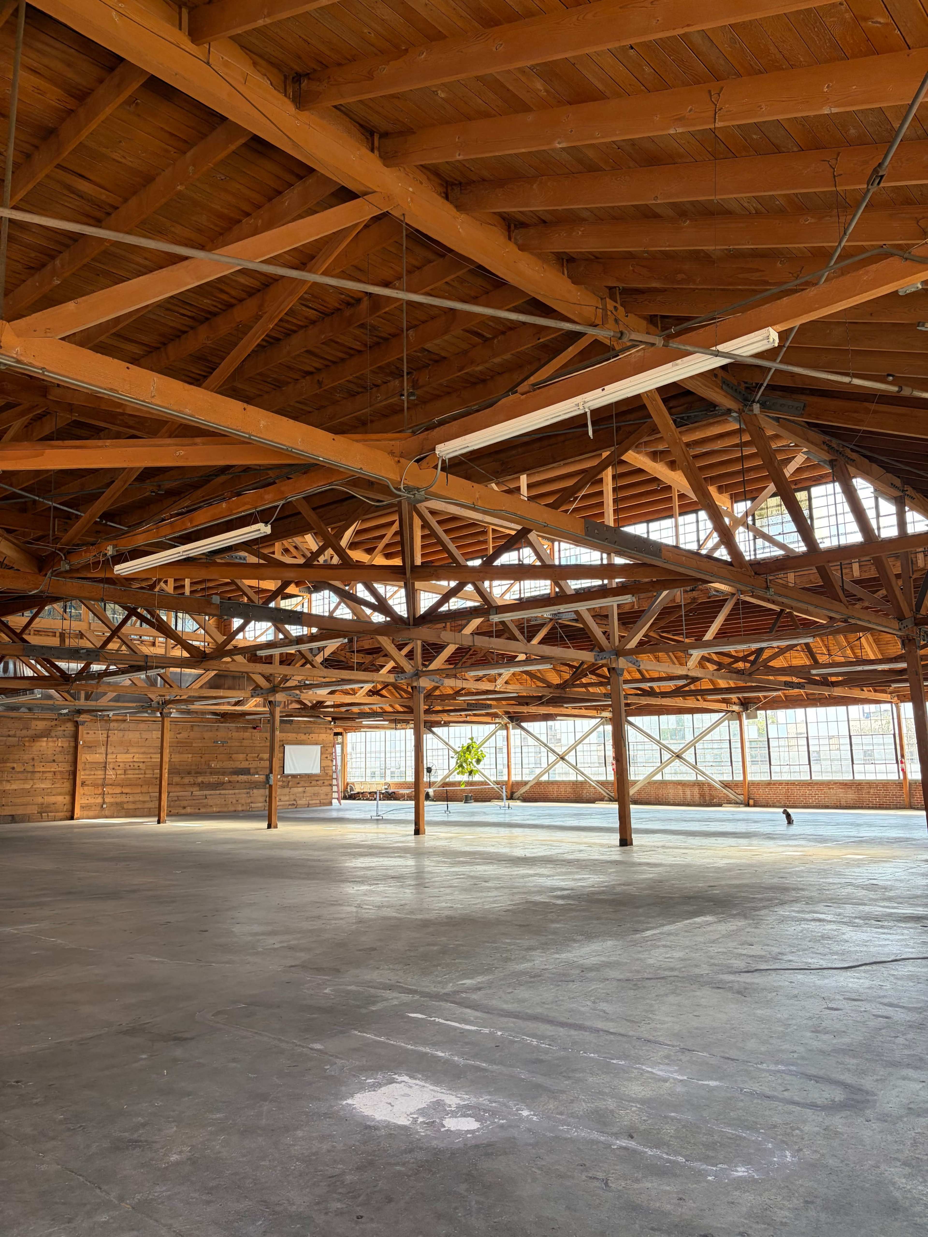 The image shows a spacious, empty warehouse with wooden beams and large windows letting in natural light.