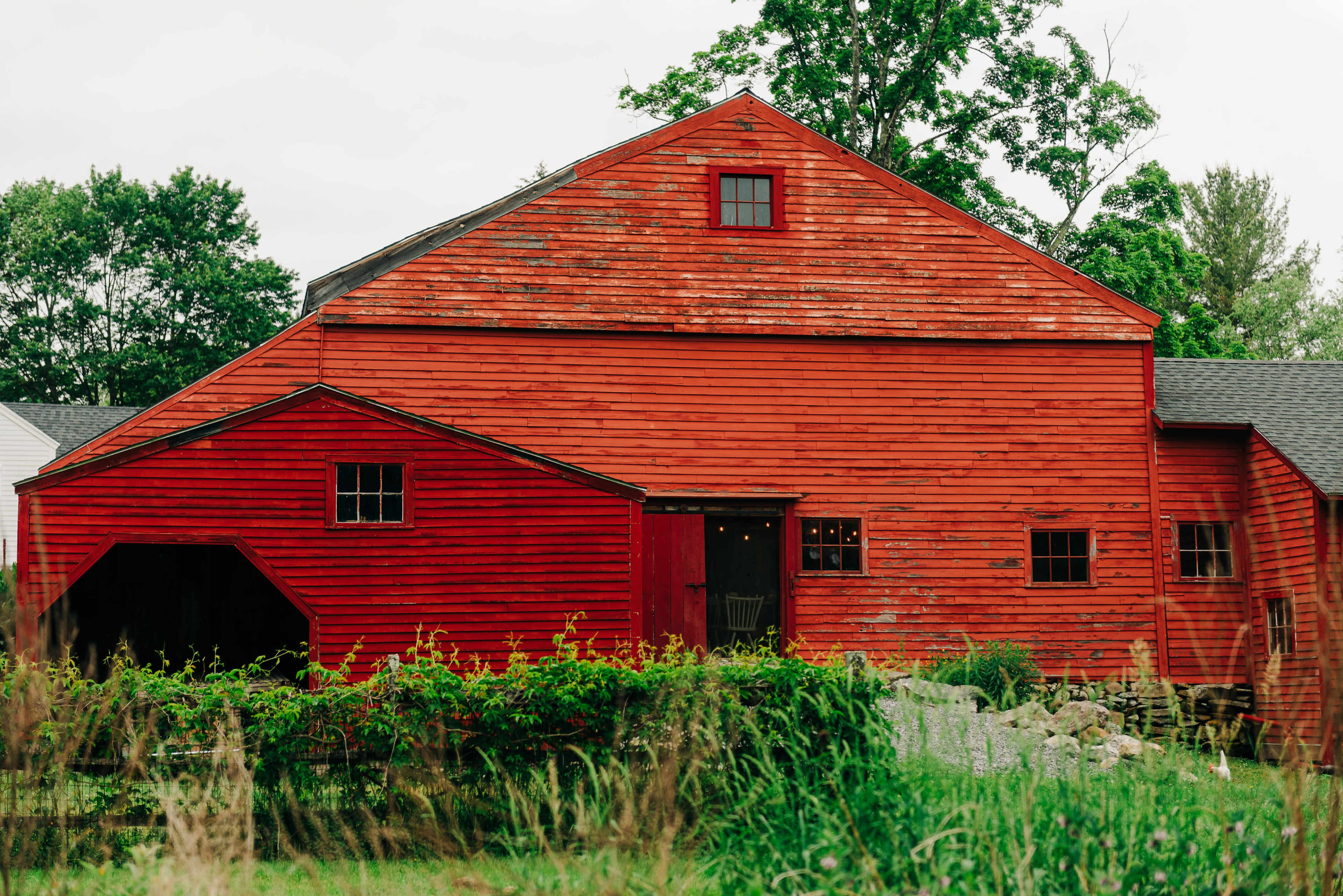A large red barn with a steep roof and multiple windows stands amidst overgrown grass and trees.