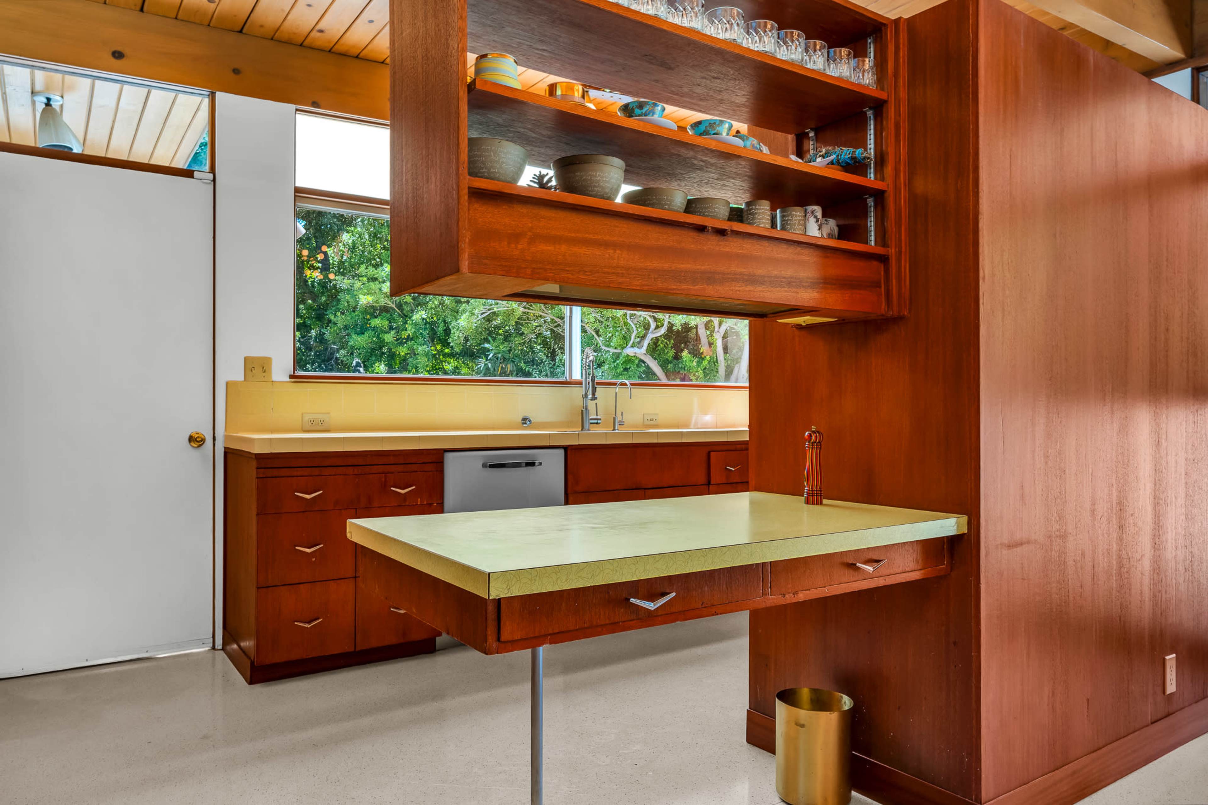 A modern kitchen features wooden cabinetry, a light green countertop, and large windows overlooking greenery.