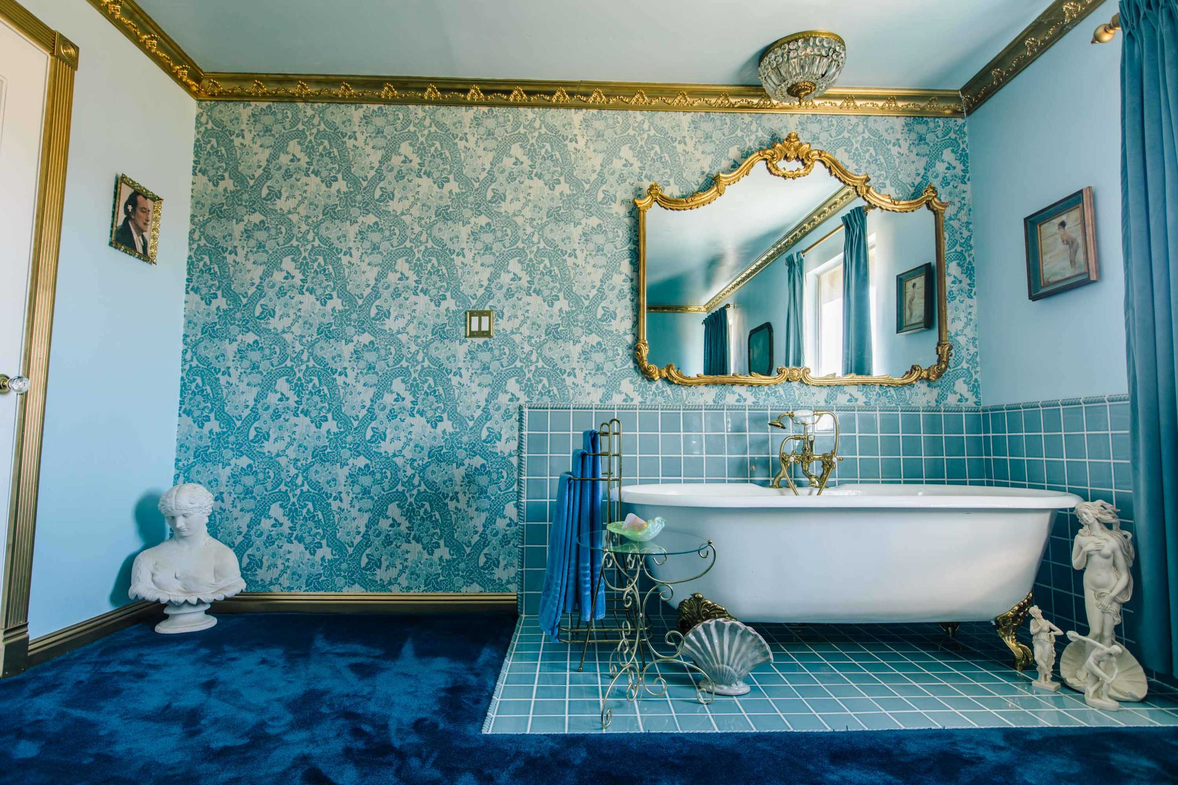 The image shows a brightly lit bathroom featuring a freestanding white bathtub, blue tiled walls, and decorative vintage elements.