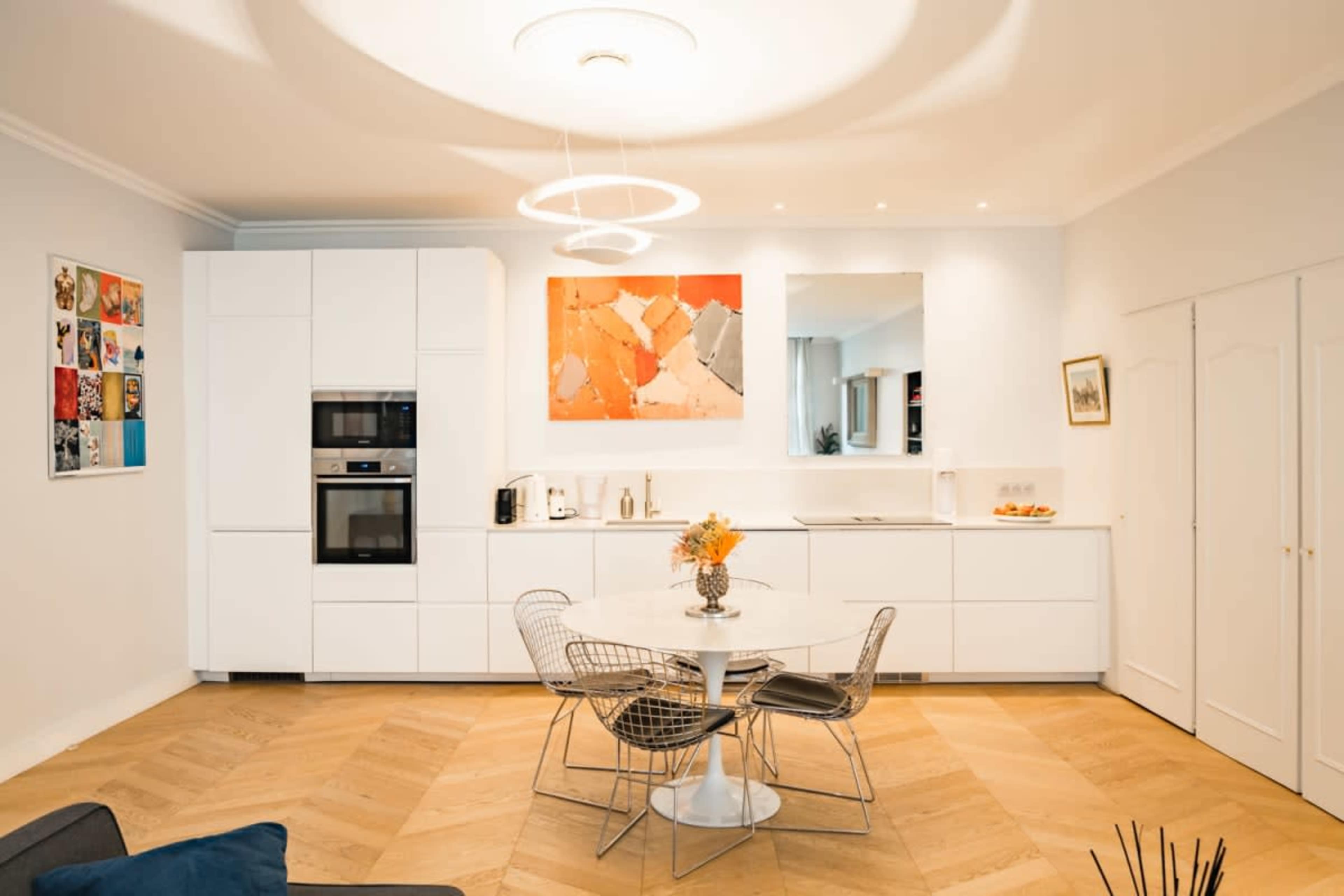 A modern kitchen and dining area featuring a round white table with metal chairs, a sleek white cabinetry setup, and artwork displayed on the walls.