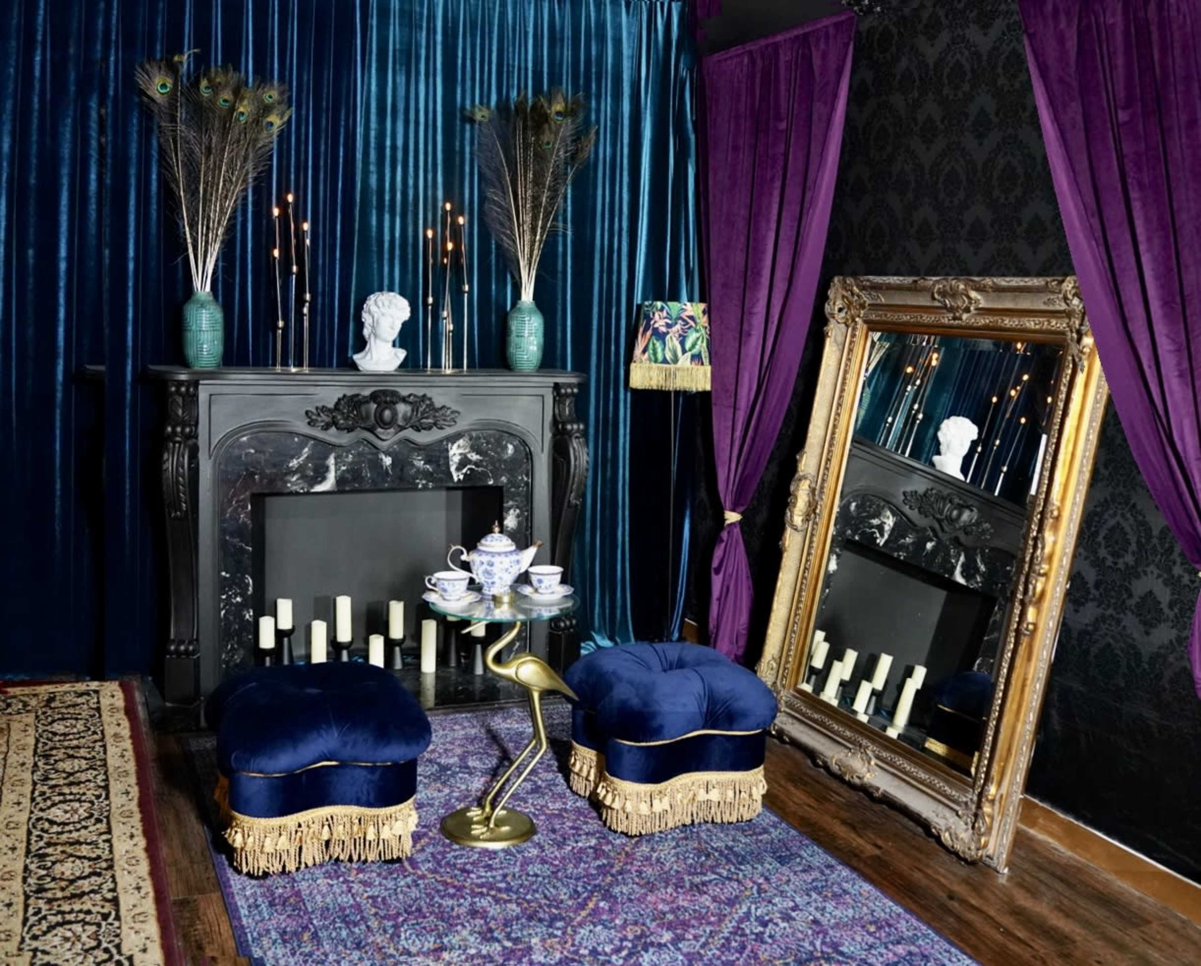 A luxurious interior scene featuring a black fireplace adorned with candles, two blue upholstered stools with fringes, a large ornate mirror, and dark velvet drapes.
