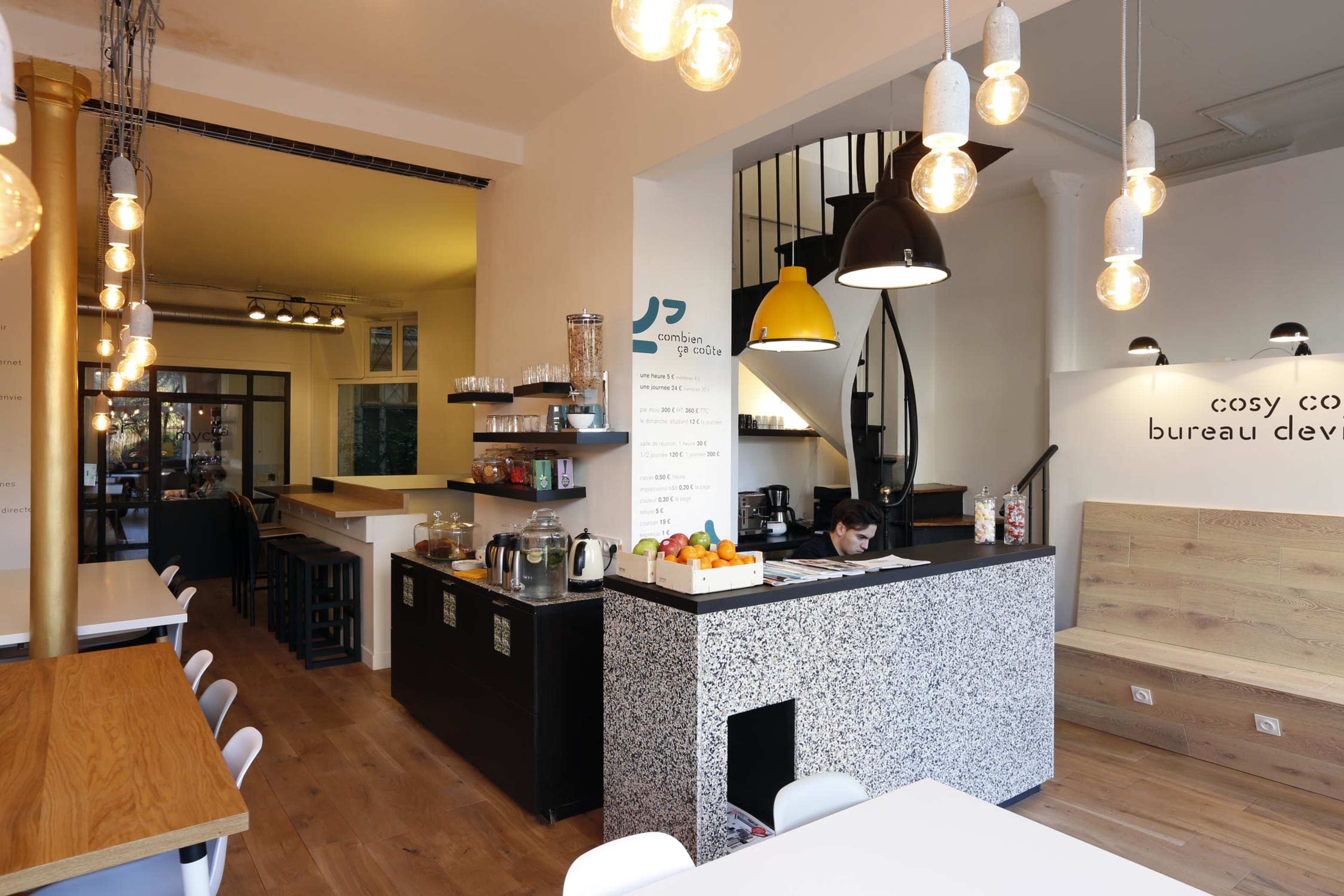 The image shows a modern café interior featuring wooden tables, a counter with a geometric design, and pendant lights hanging from the ceiling.