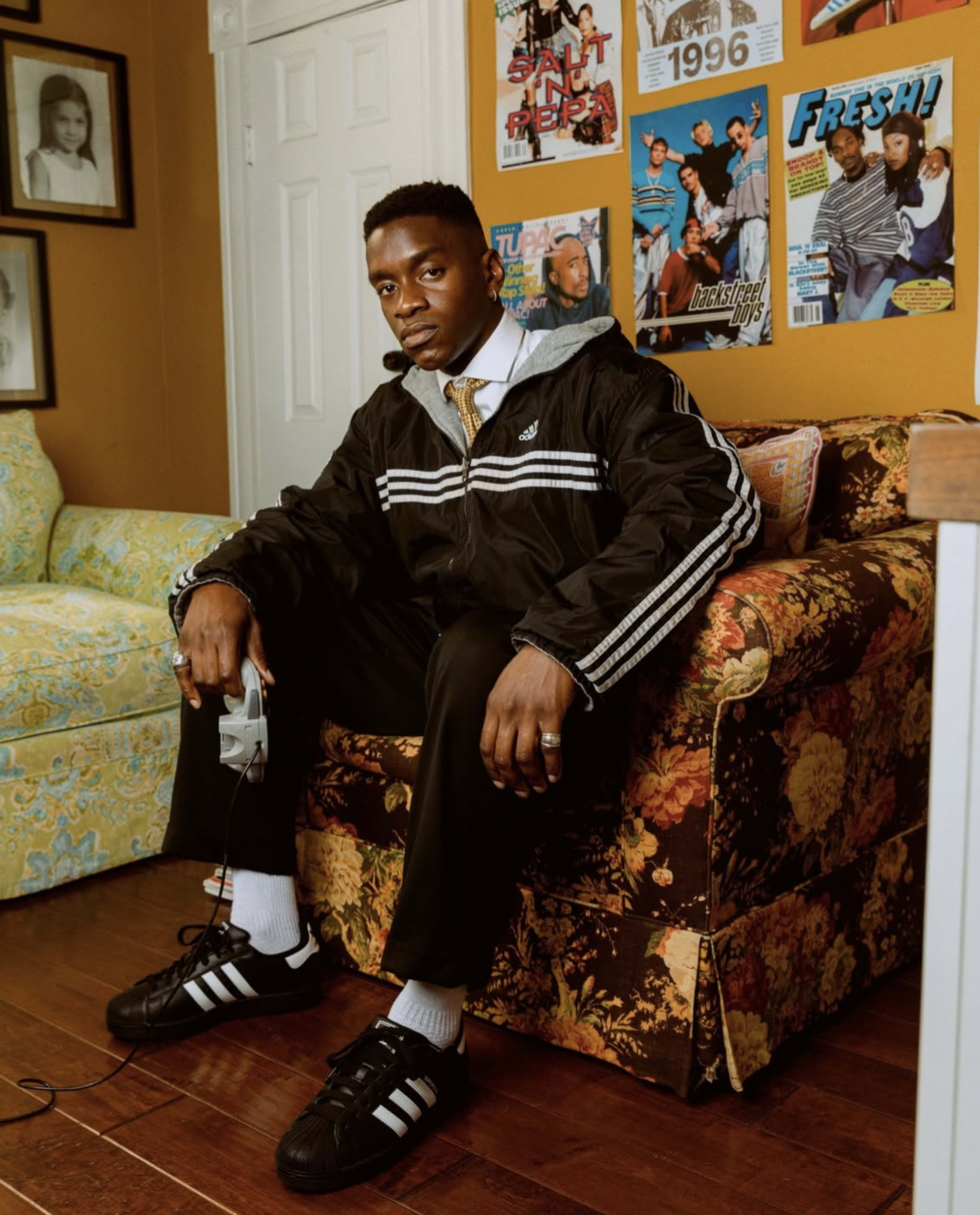 A young man wearing a black athletic jacket and pants sits on a floral-patterned sofa, holding a video game controller, with magazines and framed photos on the wall behind him.