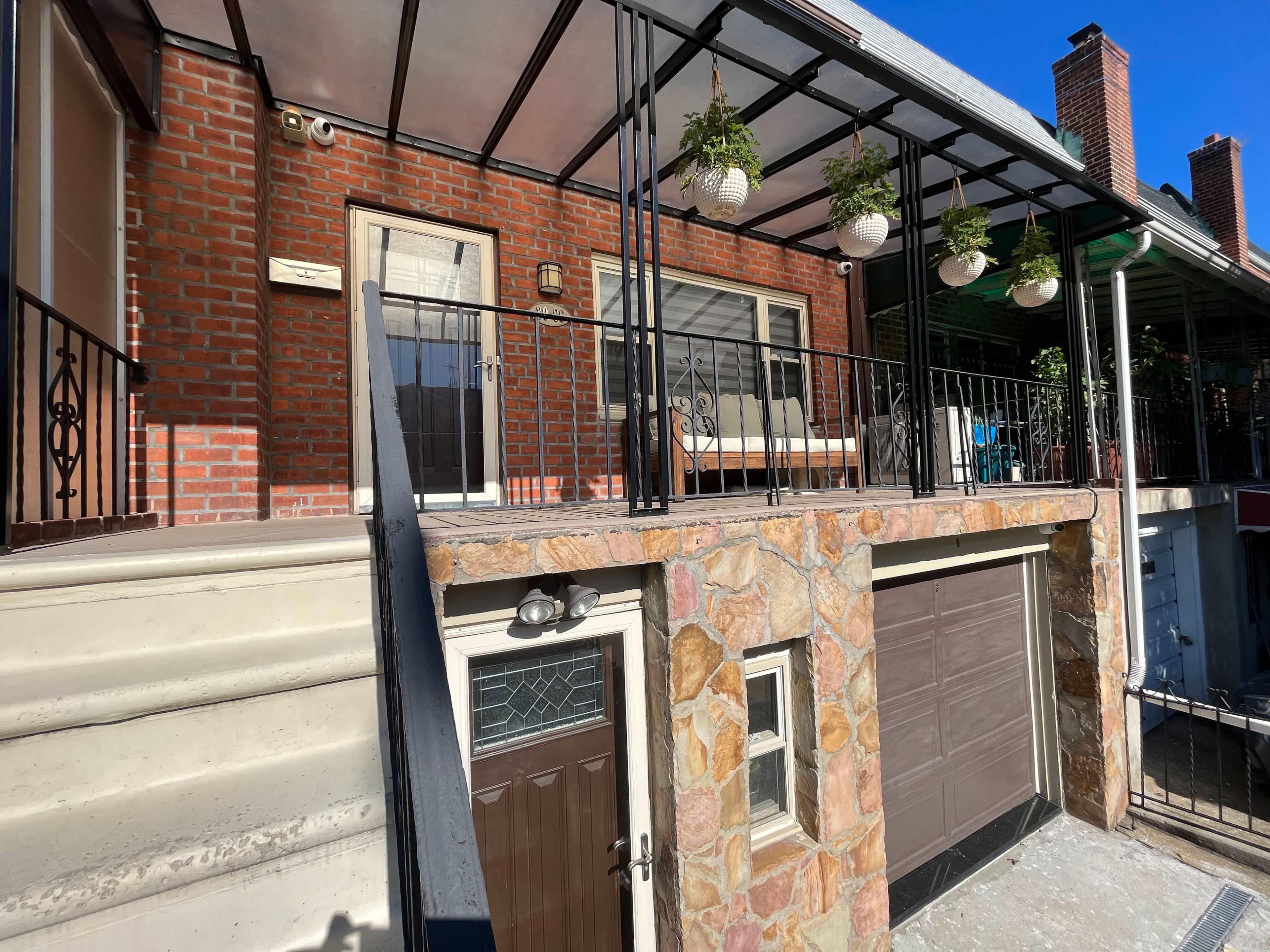 The image shows a two-story building with a brick facade, a stone wall at the garage entrance, and a balcony adorned with hanging plants.