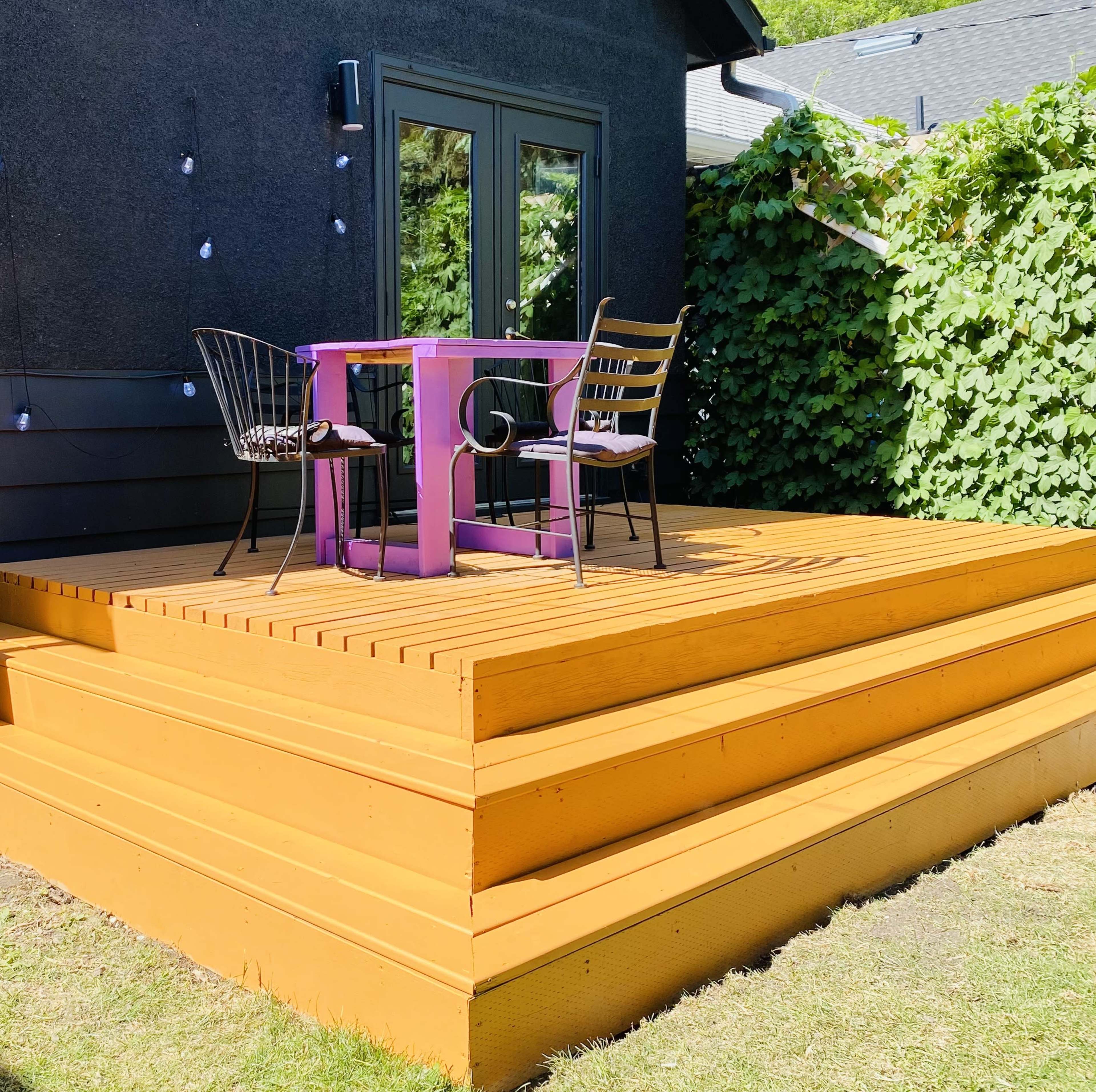 A purple table with two metal chairs is set on a wooden deck with yellow planks, surrounded by lush green foliage.