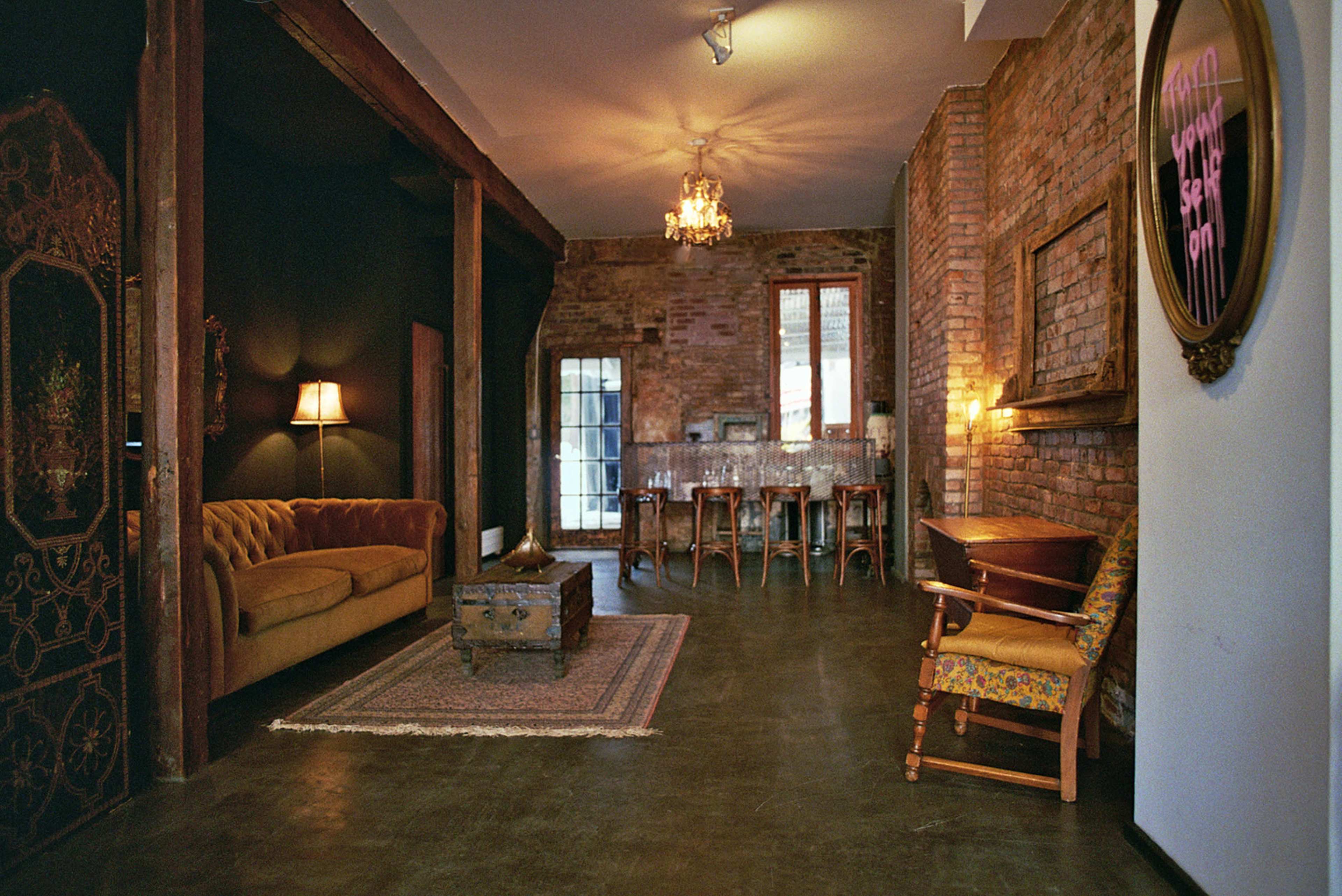 The image depicts a spacious, industrial-style living area with exposed brick walls, a vintage sofa, and a dining area featuring a large table and elegant lighting.