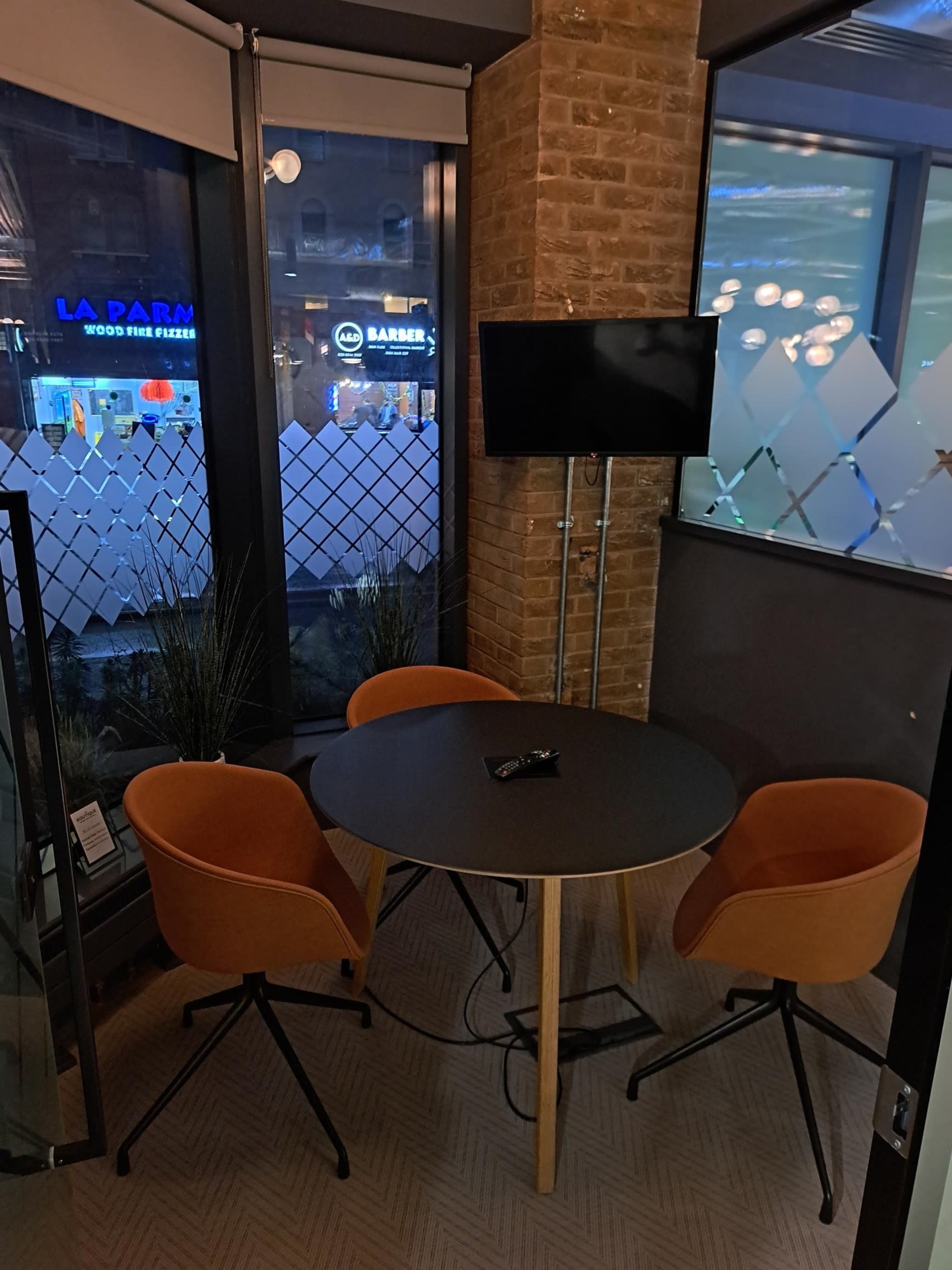 A small circular table with two orange chairs and a wall-mounted TV in a corner office space with large windows.