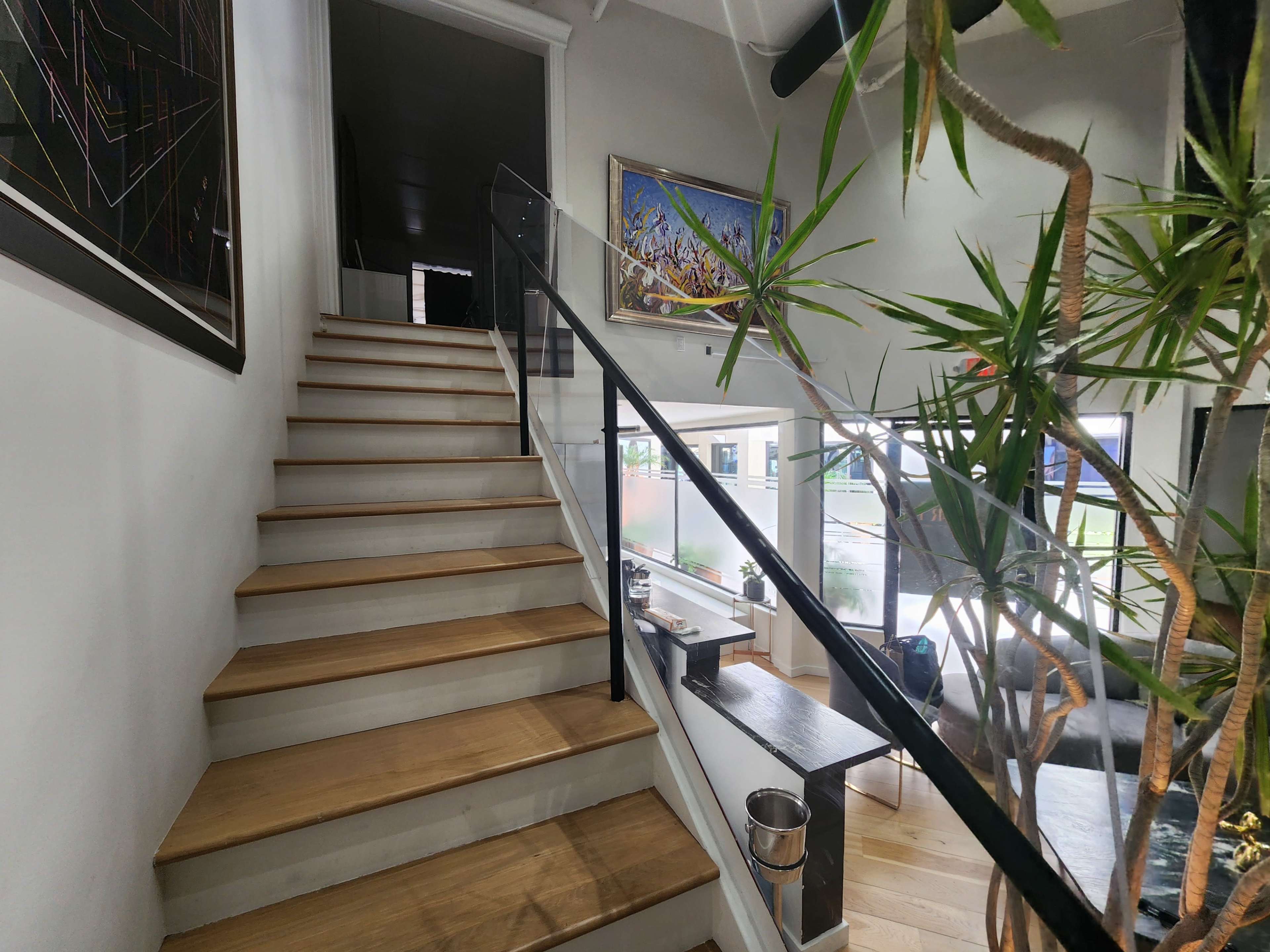 The image shows a wooden staircase leading to an upper level, flanked by potted plants and featuring a glass railing, with an open living area visible below.