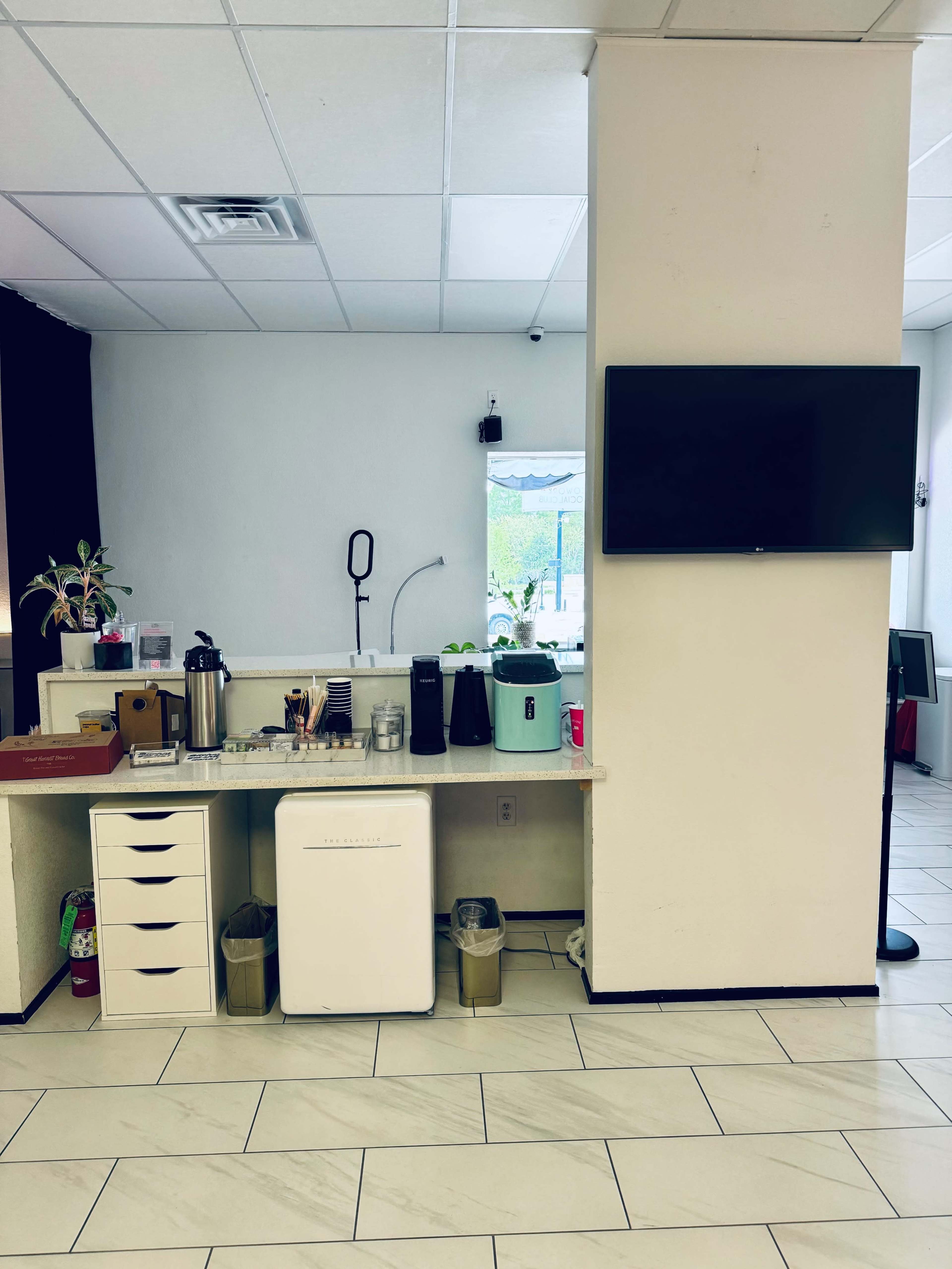 The image shows a small service area with a countertop featuring a coffee machine, several containers, and a refrigerator, alongside a television mounted on the wall.