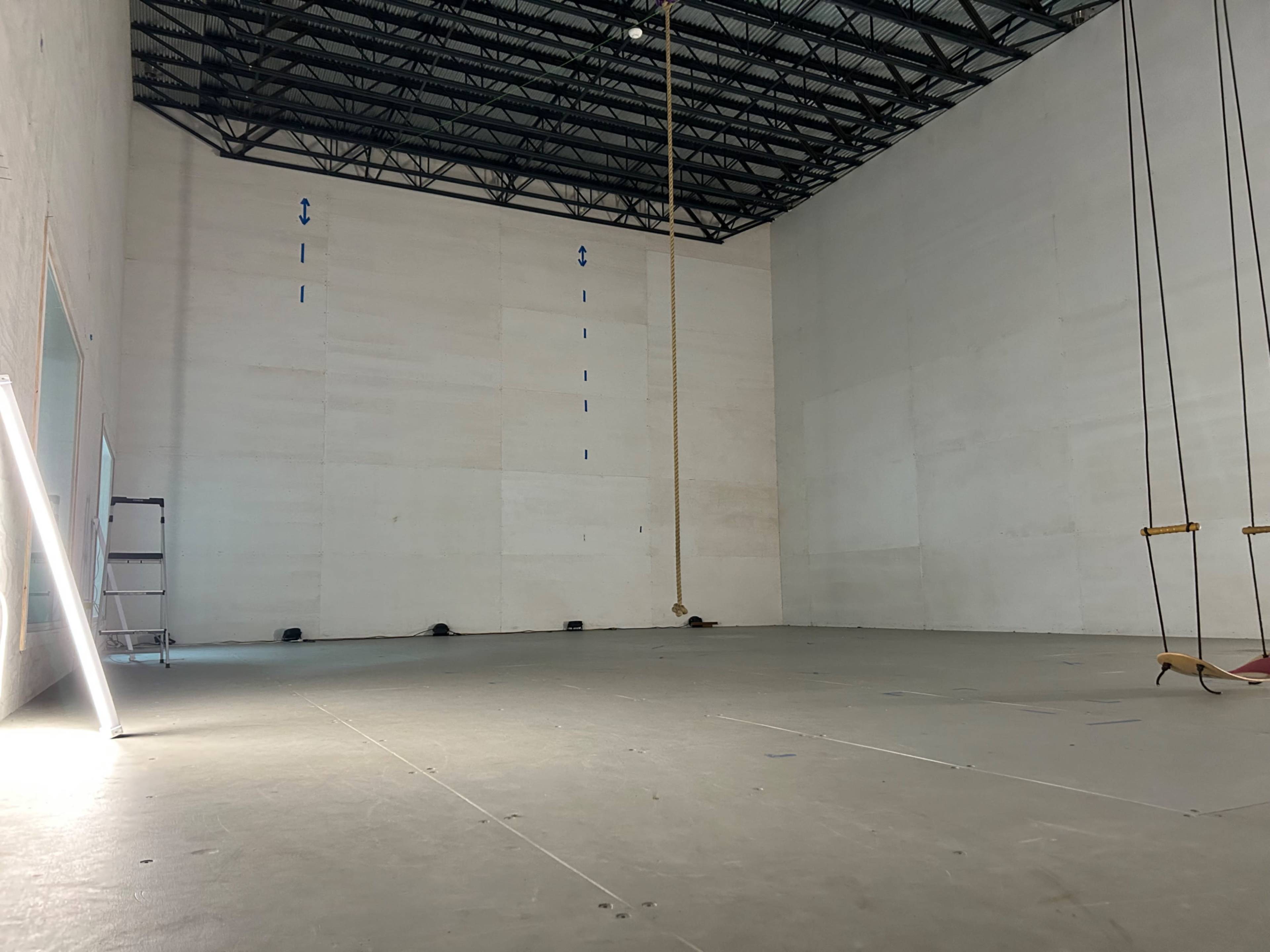 A spacious, empty room with a high ceiling, a rope hanging from the ceiling, and a ladder positioned against the wall.