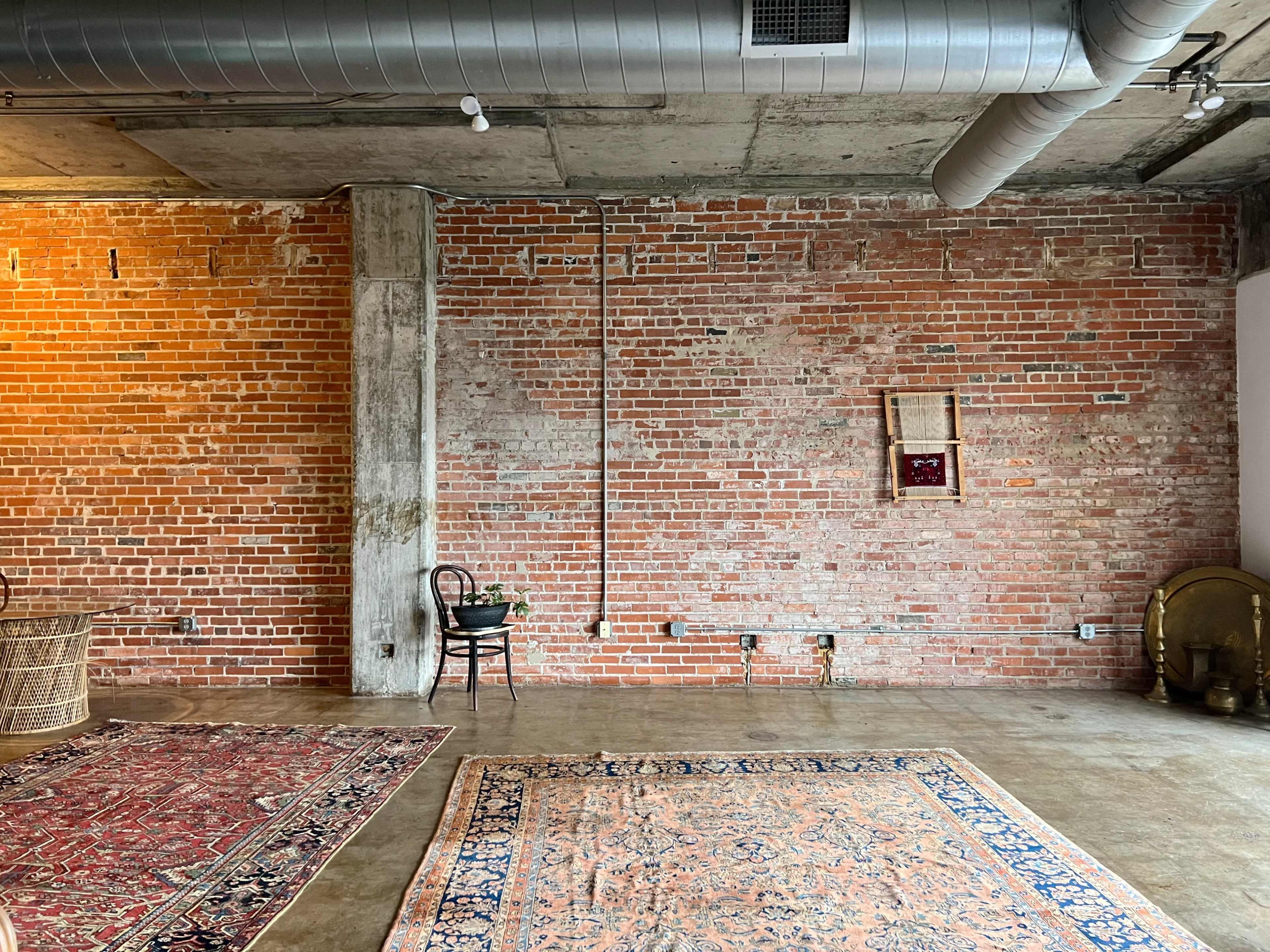The image shows a spacious room with exposed brick walls, a concrete floor, and various rugs scattered across the area.