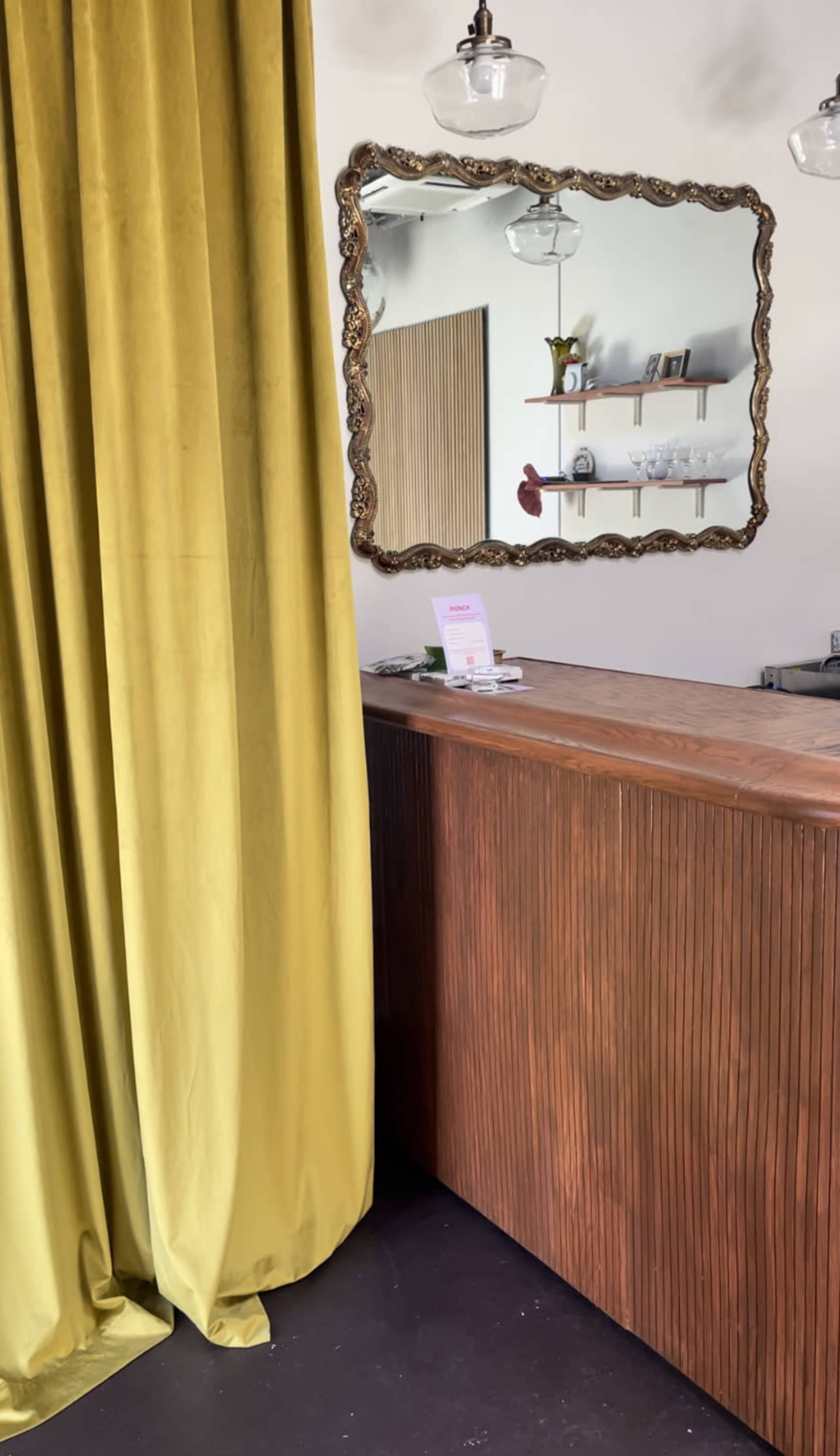 A wooden reception desk next to a long yellow curtain and a decorative mirror on the wall, with shelves displaying items above the desk.