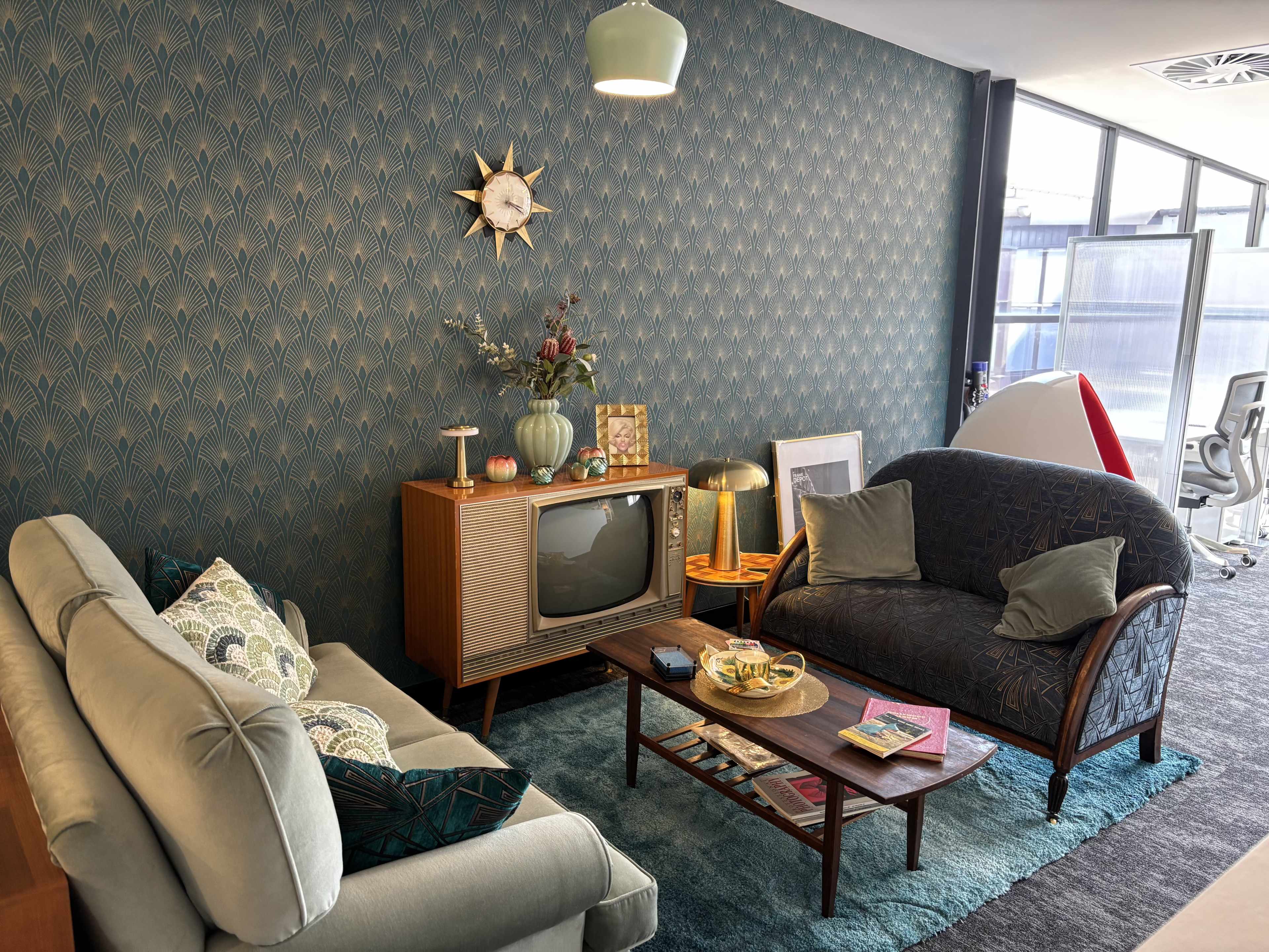 The image shows a mid-century modern living room with two sofas, a vintage television, a coffee table, and decorative elements against a patterned wallpaper backdrop.