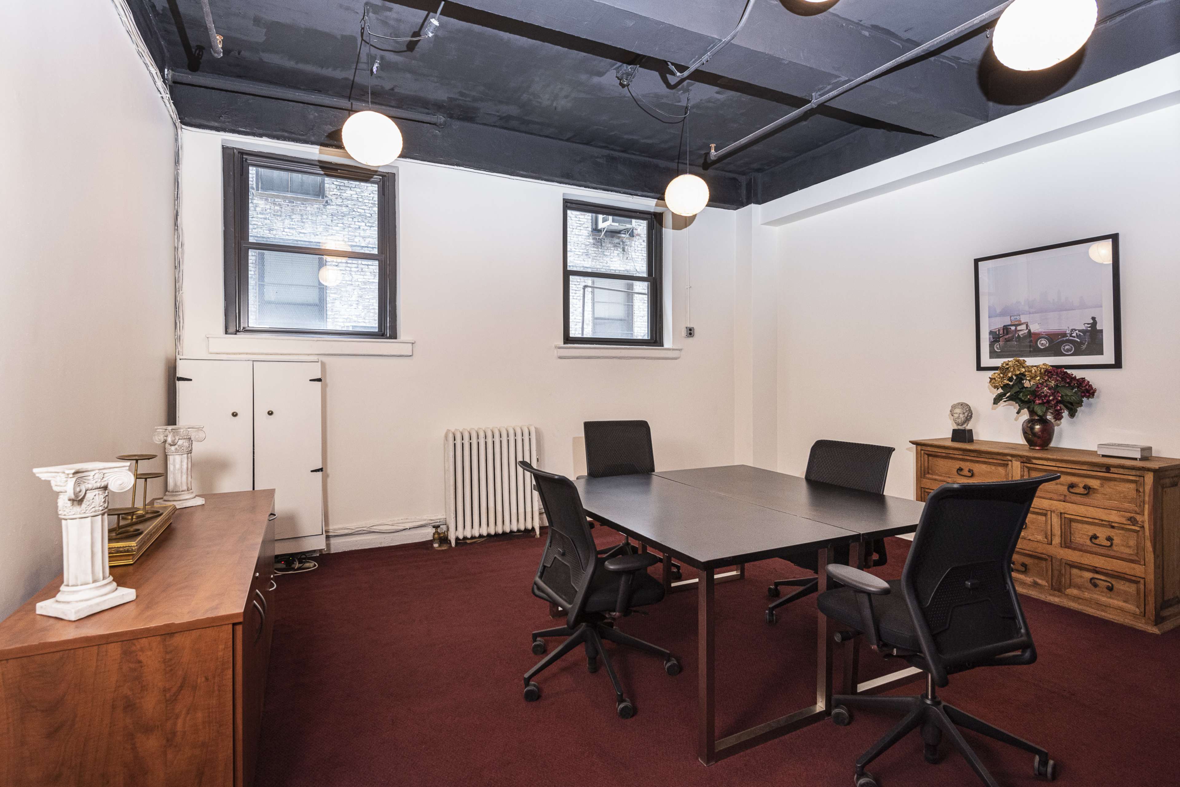 A conference room features a rectangular table surrounded by four chairs, with windows, a cabinet, and a decorative photo on the wall.