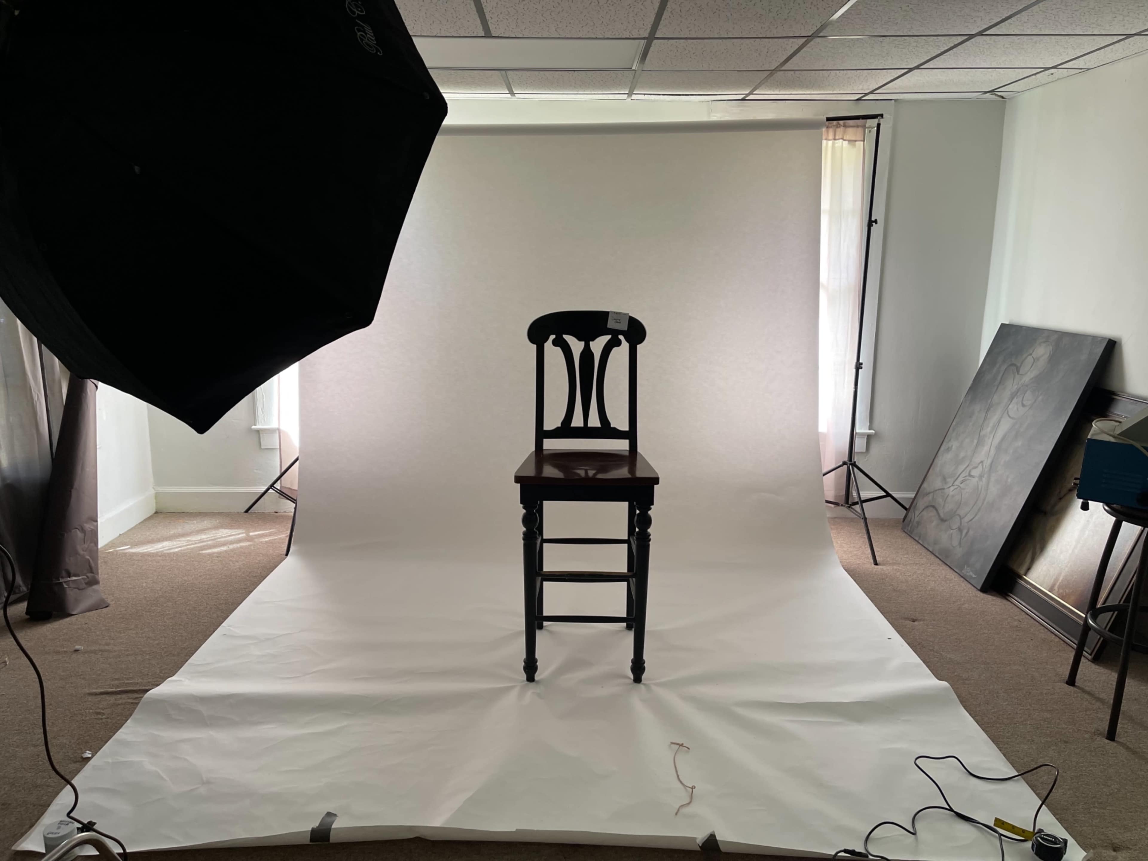 A black chair is centered in a brightly lit studio surrounded by white backdrop paper and photography equipment.