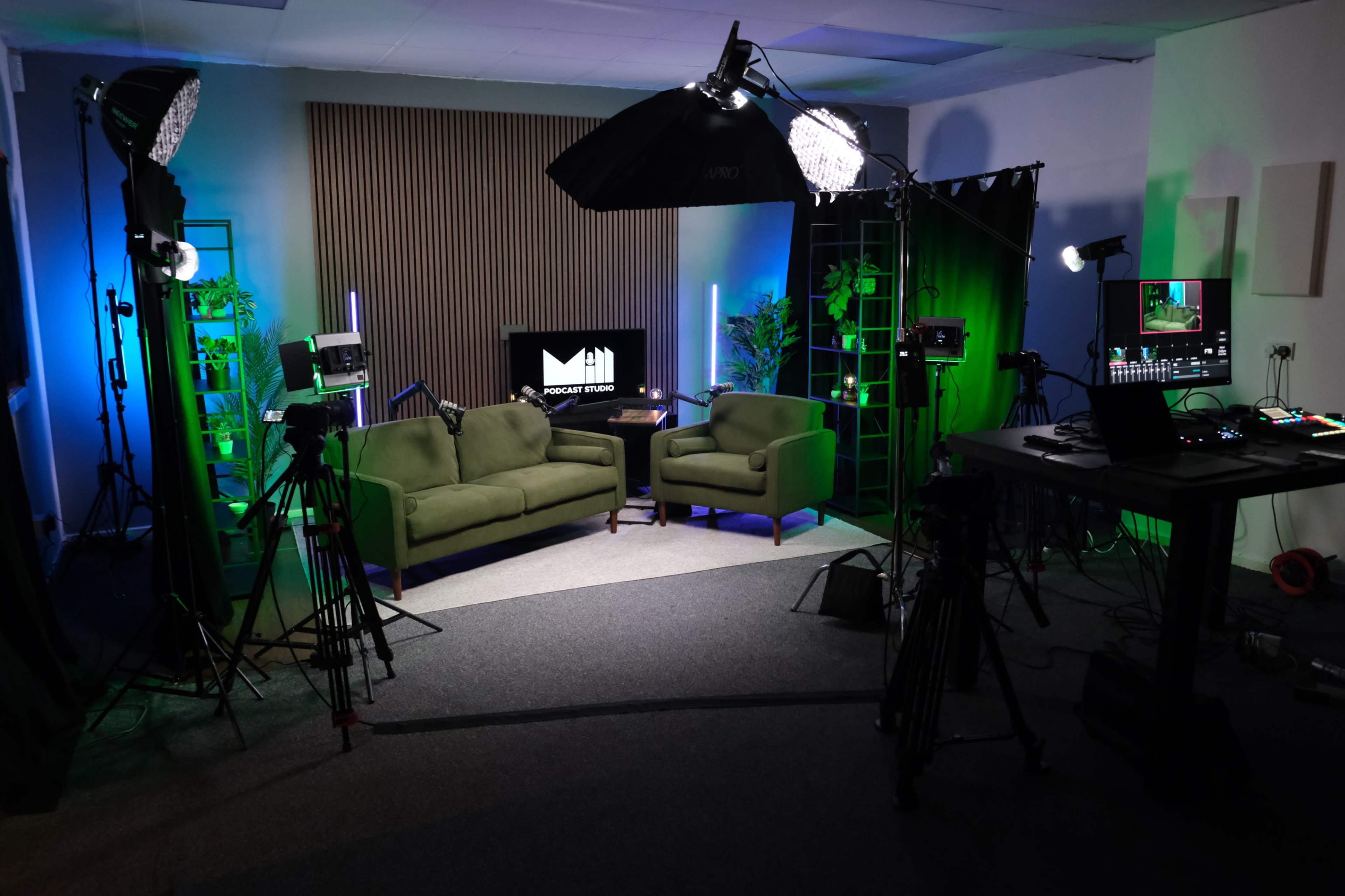 A well-equipped podcast studio features two green couches, multiple cameras, lighting setups, and a monitor displaying the project logo.