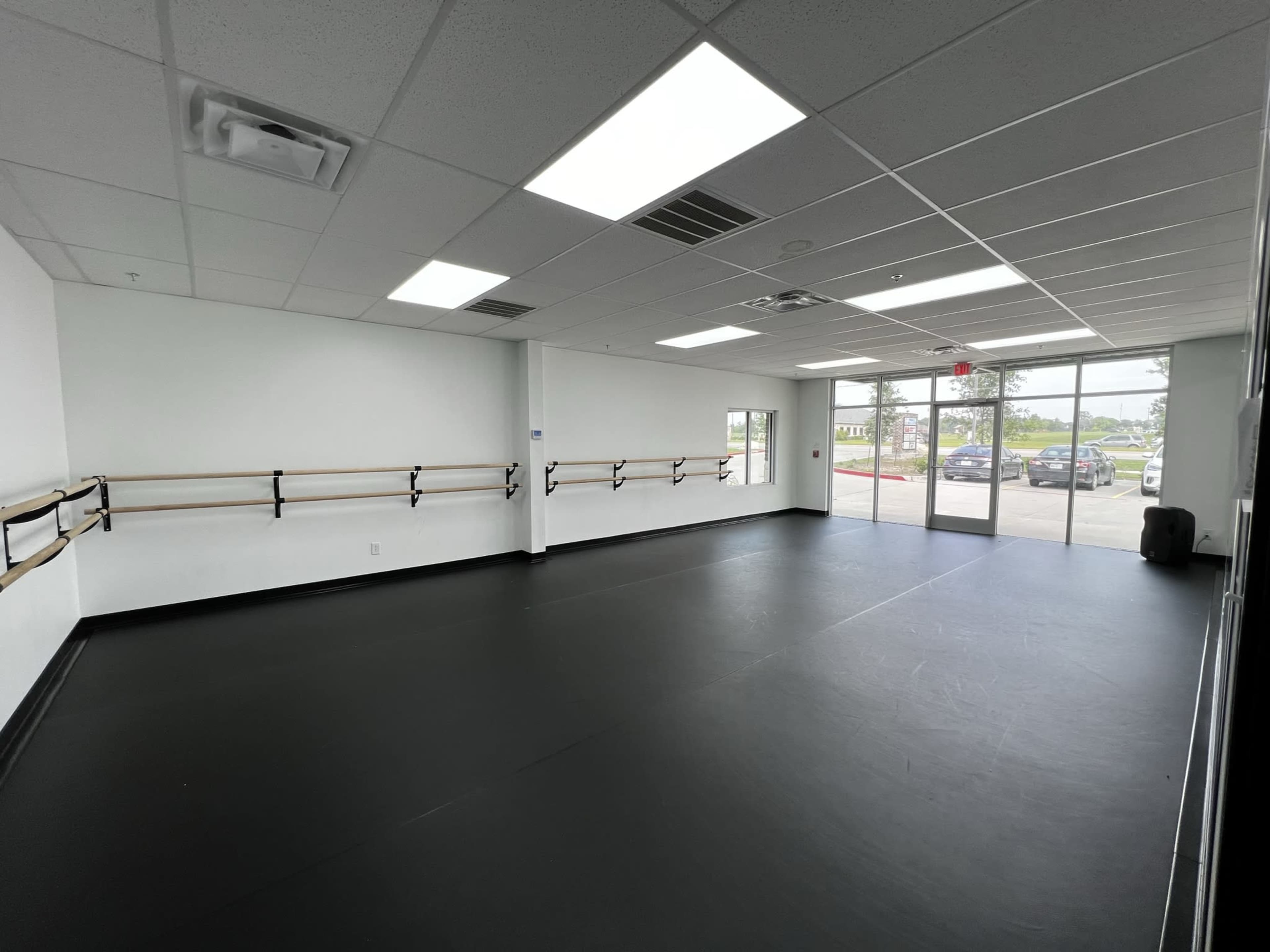 A spacious dance studio with a black floor, mirrored walls, and ballet barres along one side, along with large windows letting in natural light.
